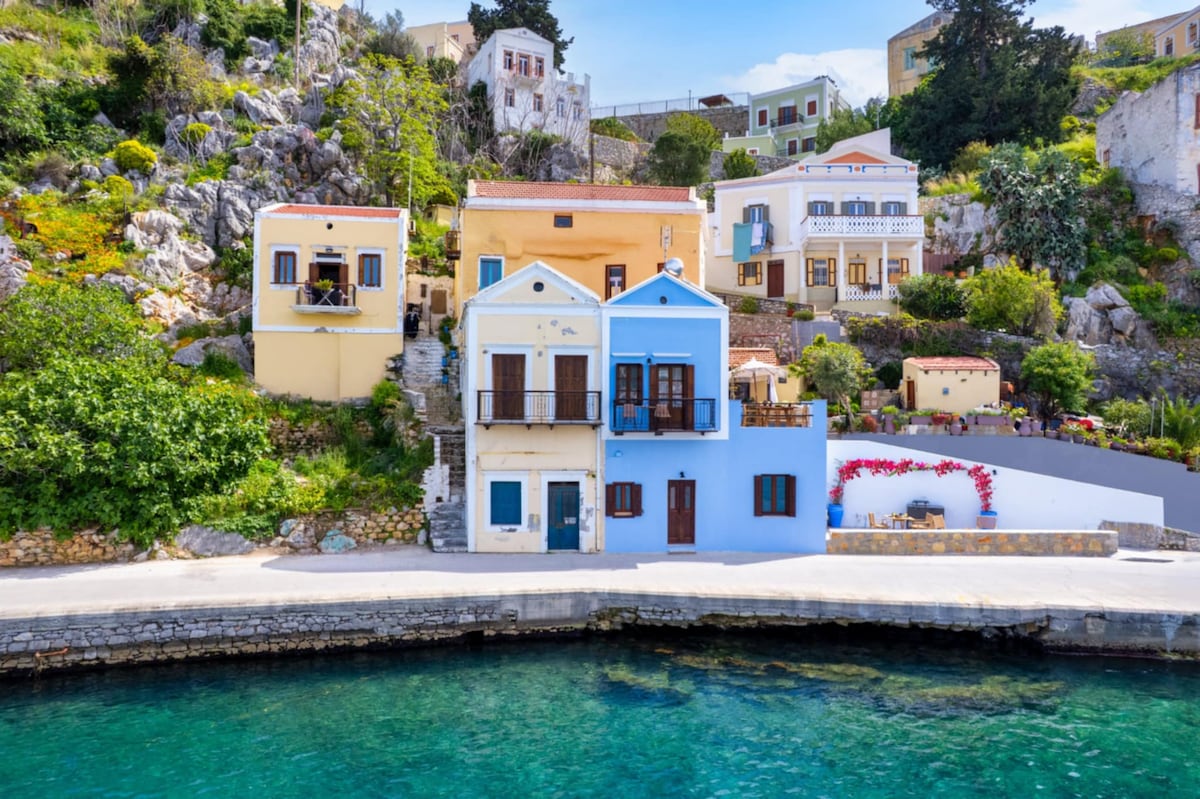A collection of traditional houses is positioned along the shoreline, displaying a range of pastel colors. Lush greenery and rocky terrain serve as a backdrop. The tranquil waters of Gialos harbor reflect the vibrant shades of the structures, enhancing the charm of this coastal setting.