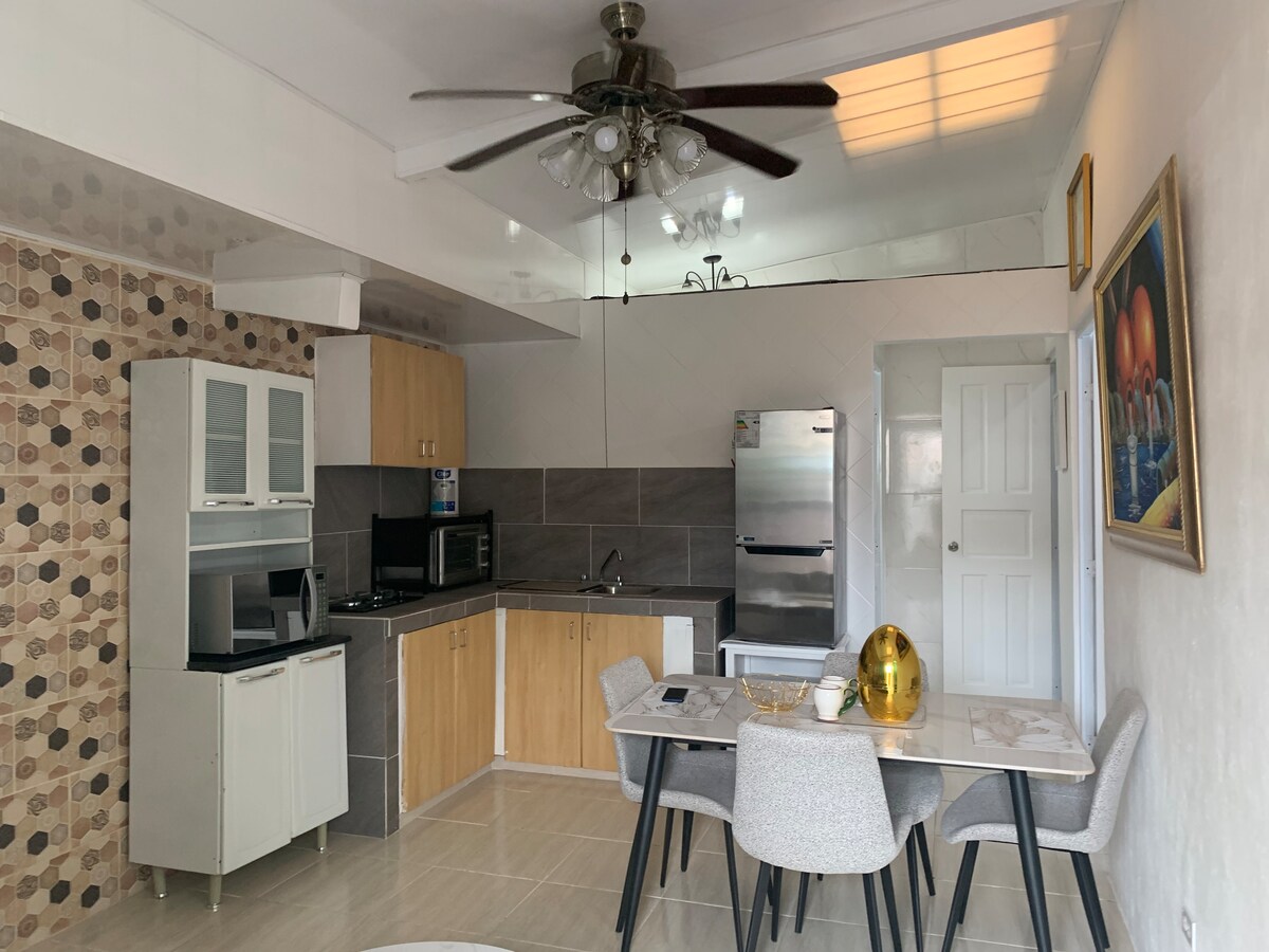 An open kitchen and dining area is visible, featuring wooden cabinets and modern appliances. A dining table, surrounded by upholstered chairs, is positioned near a large window. Neutral tones and a patterned accent wall contribute to the spacious ambiance.