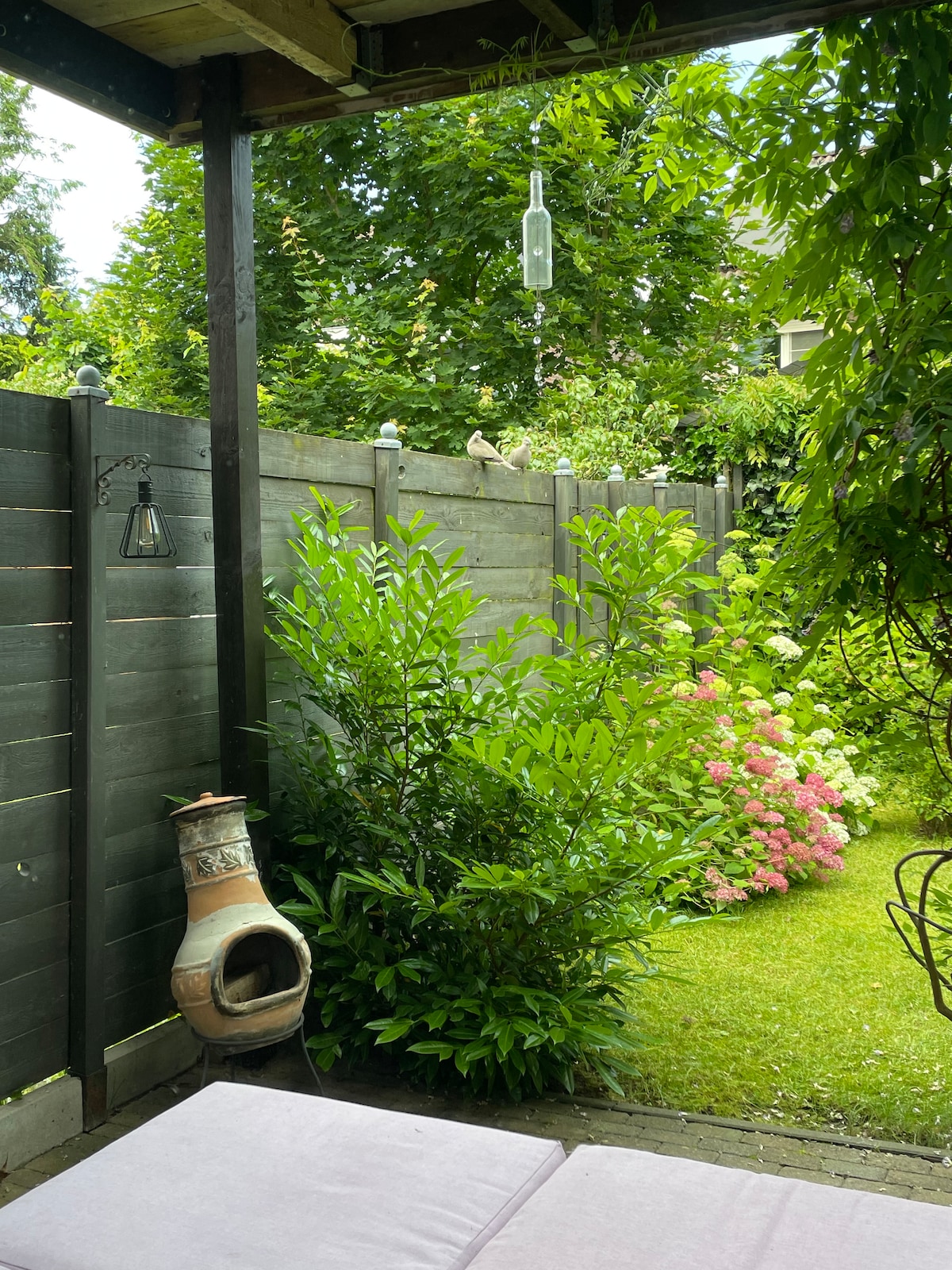 A green and inviting outdoor space is surrounded by a wooden fence, featuring lush plants and flowering shrubs. A small, decorative chiminea stands beside a comfortable seating area, with natural light filtering through the foliage above, creating a serene atmosphere.