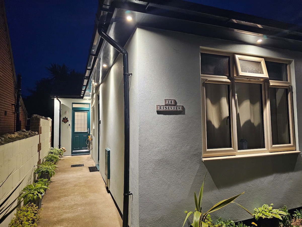 The exterior of the accommodation is presented at night, featuring clean lines and soft lighting illuminating the entrance. A welcoming blue door is complemented by large windows adorned with curtains. Potted plants are positioned along the pathway, enhancing the inviting appearance of the property.
