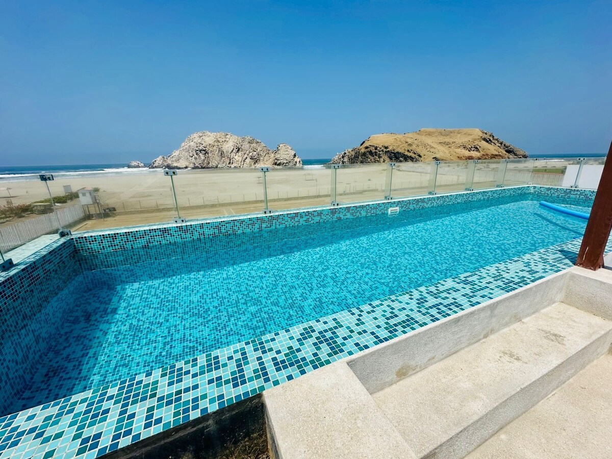 A large swimming pool sits on a third-floor terrace, showcasing vibrant blue tiles. The view beyond features the ocean and a distant rocky island, framed by a clear blue sky. Glass railings surround the pool area, offering a sense of openness.