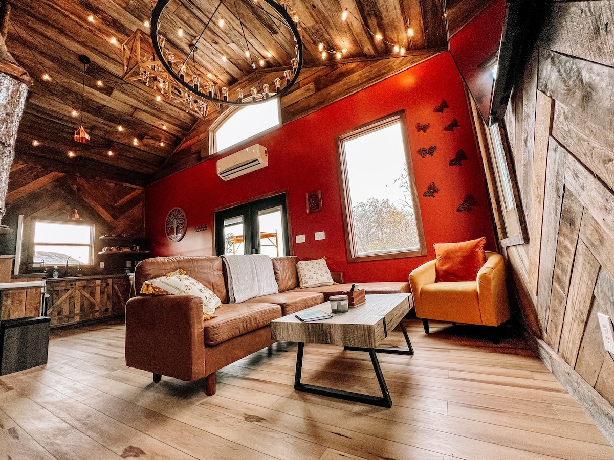 A warm living area features a leather sofa and an orange accent chair, complemented by a rustic wooden coffee table. Natural light streams through the large windows, highlighting the textured wooden walls and ceiling adorned with string lights.