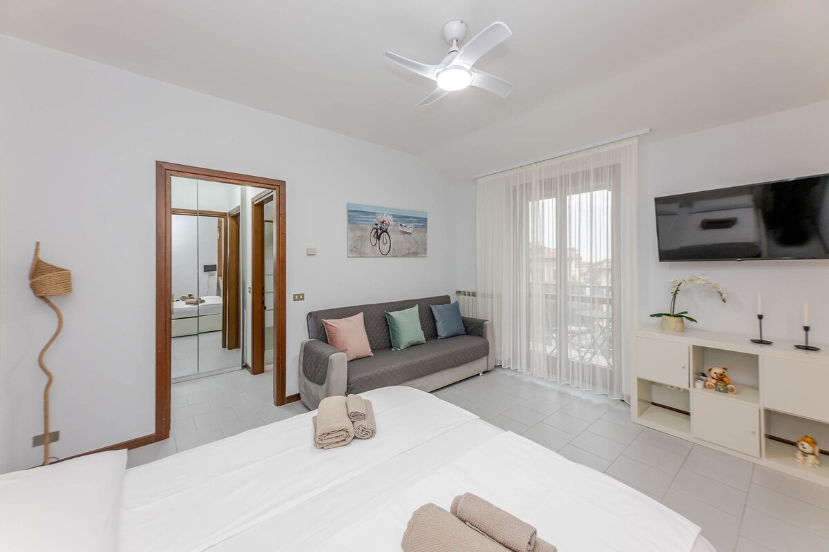 An open space featuring a large bed with fresh linens, complemented by a cozy sofa against the wall. Natural light filters through the sheer curtains, illuminating the room. A ceiling fan enhances comfort, while a modern TV and tasteful decor complete the inviting atmosphere.