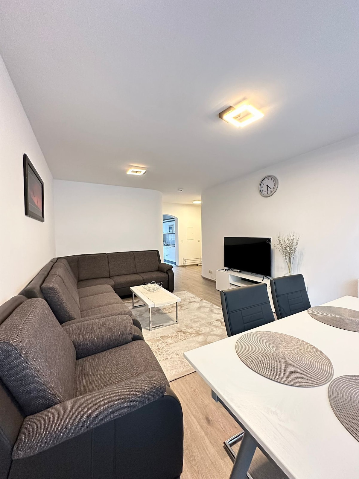 A modern living area is observed, featuring a spacious sectional sofa and a coffee table. A dining area with sleek black chairs is present, alongside a white table. A television is positioned against the wall, with a decorative clock hanging above. Natural light enhances the neutral color palette.