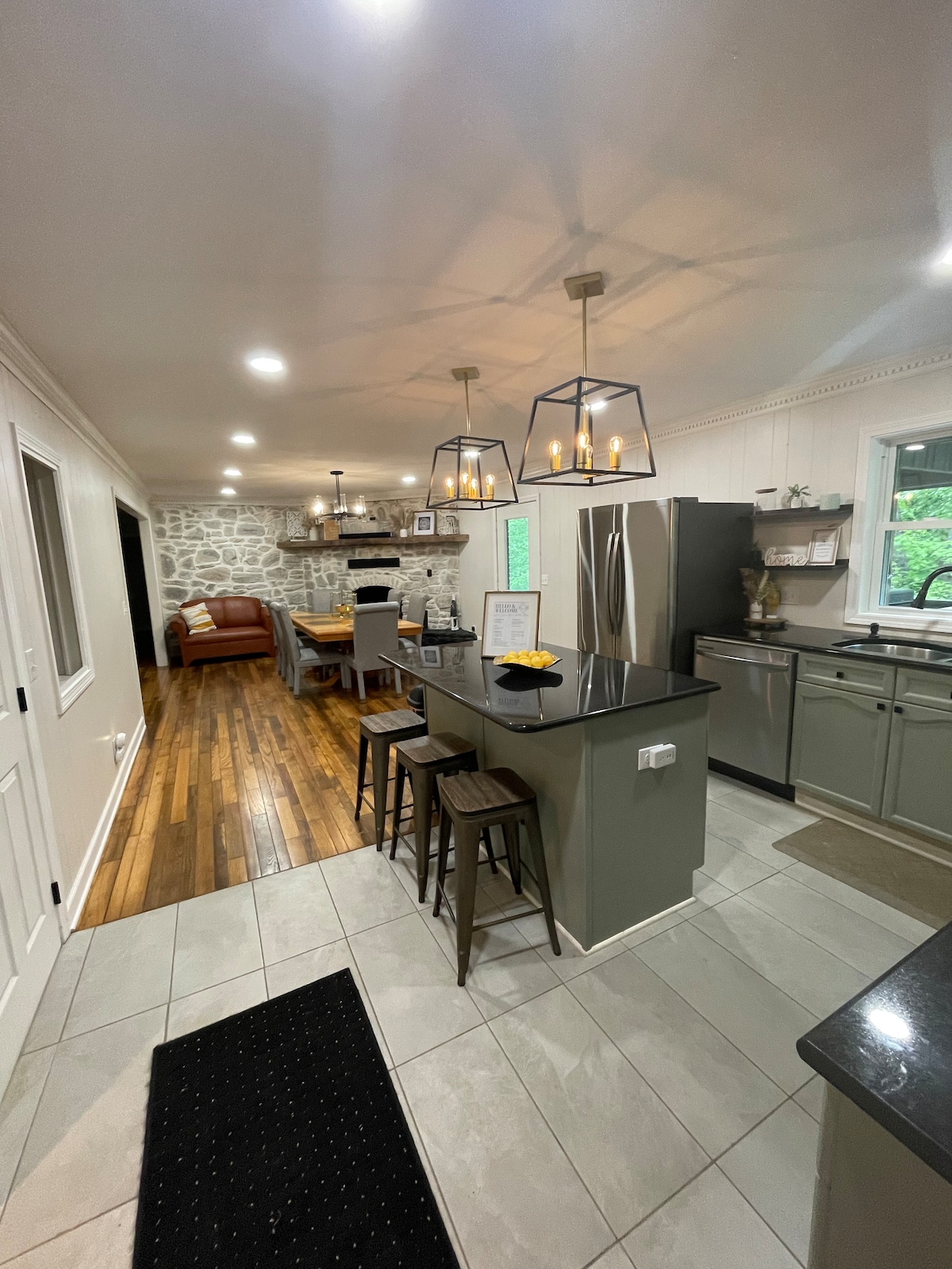 An open kitchen with dark countertops is complemented by light cabinetry and modern fixtures. Three bar stools are positioned along a kitchen island. A cozy living area is visible in the background, featuring a leather couch and stone accent wall. Natural light floods the space.