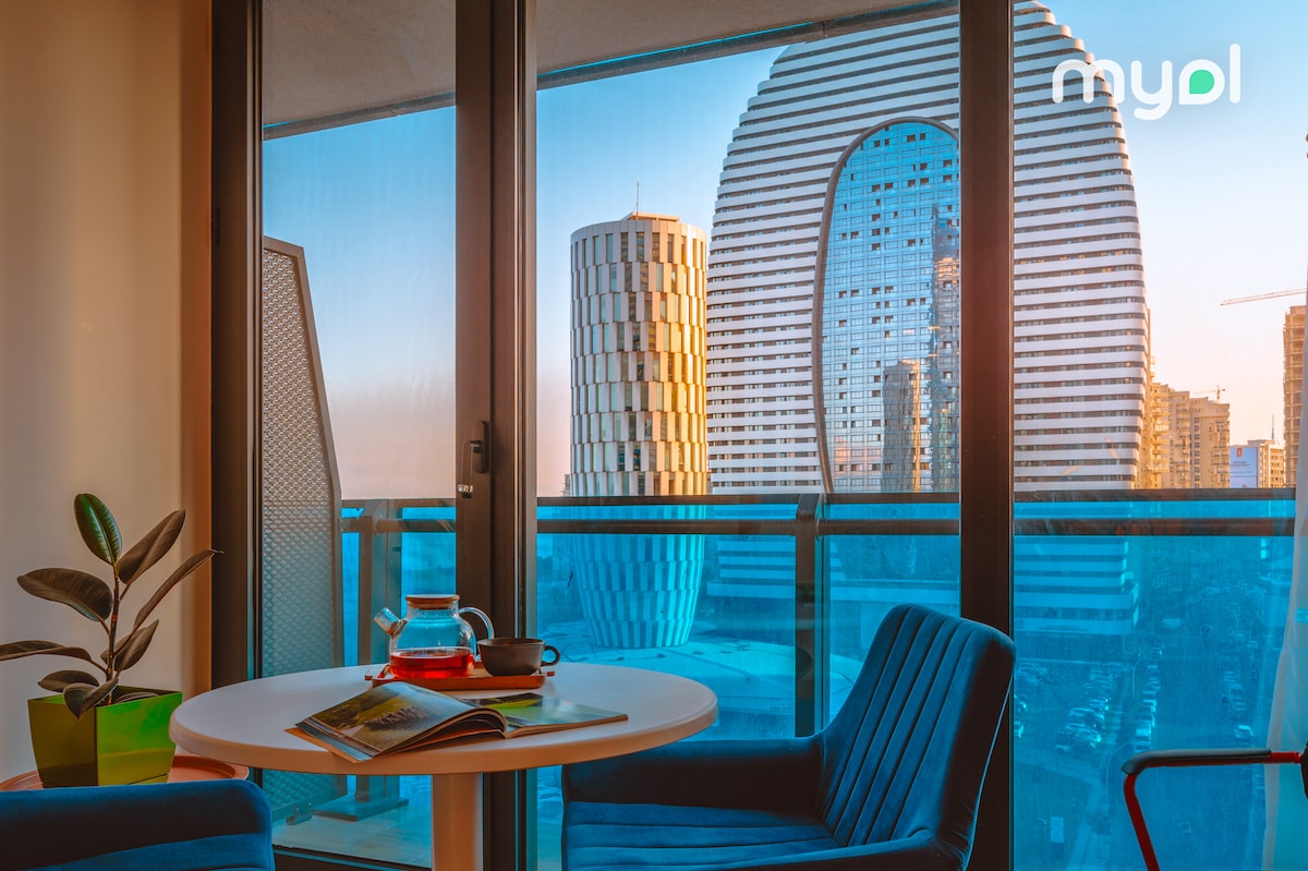 A modern balcony view showcases a circular table with a teapot and a book. Lush green plants are present, while large windows frame a striking urban skyline featuring uniquely shaped buildings and a vibrant blue sky.