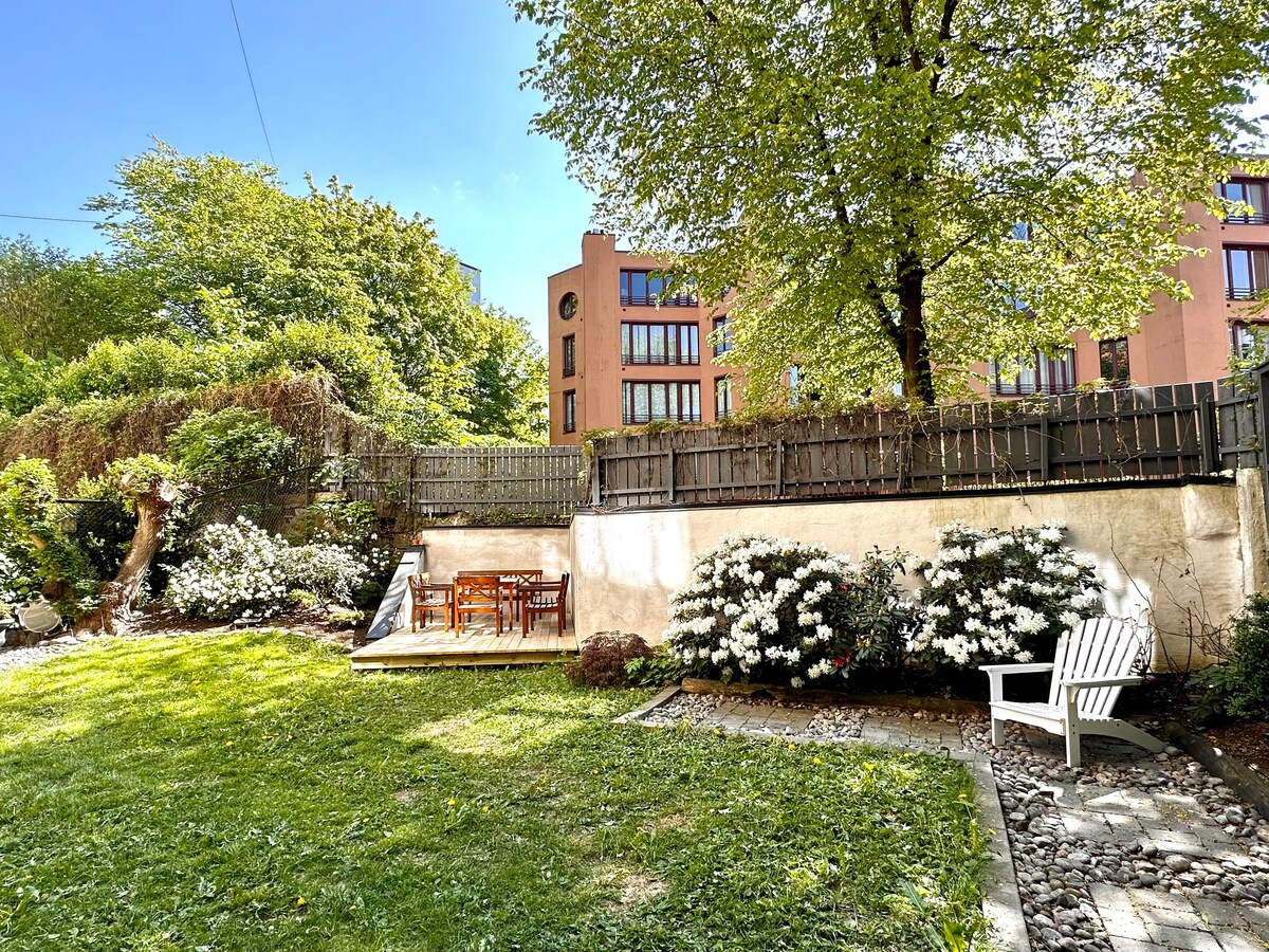 A tranquil outdoor space is highlighted by a well-kept lawn, surrounded by lush greenery and flowering bushes. A wooden dining set is situated on a patio area, with a single white lounge chair spaced comfortably on the grass. Sunlight bathes the scene under a clear blue sky.