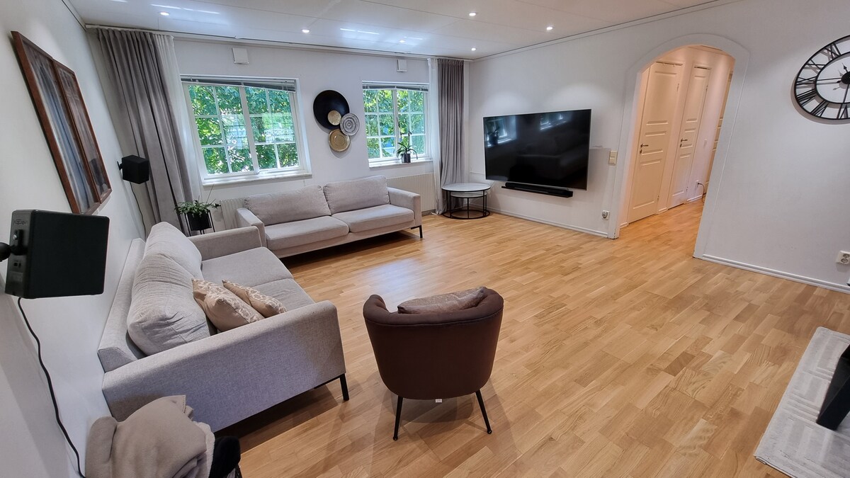 A spacious living room features two light grey sofas and a brown armchair, arranged on polished wooden flooring. Large windows allow natural light to enter, accompanied by neutral-colored curtains. A wall-mounted television is positioned opposite the seating area, complemented by a wall clock and decorative elements.
