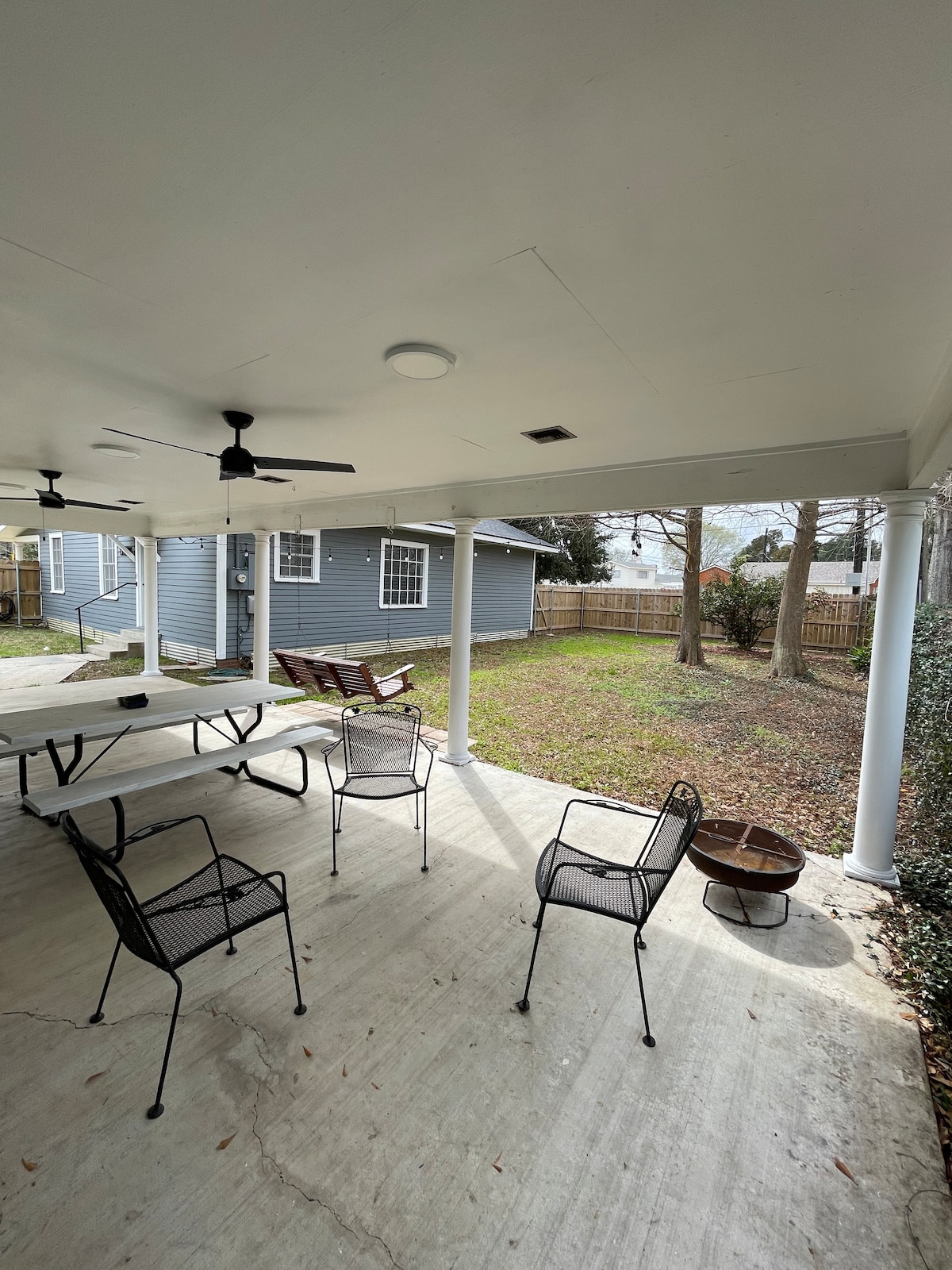 An outdoor patio area is depicted, featuring a picnic table and four metal chairs arranged around a small fire pit. The space is covered with a roof and is adjacent to a grassy yard, showcasing a serene environment. Two ceiling fans are installed overhead.