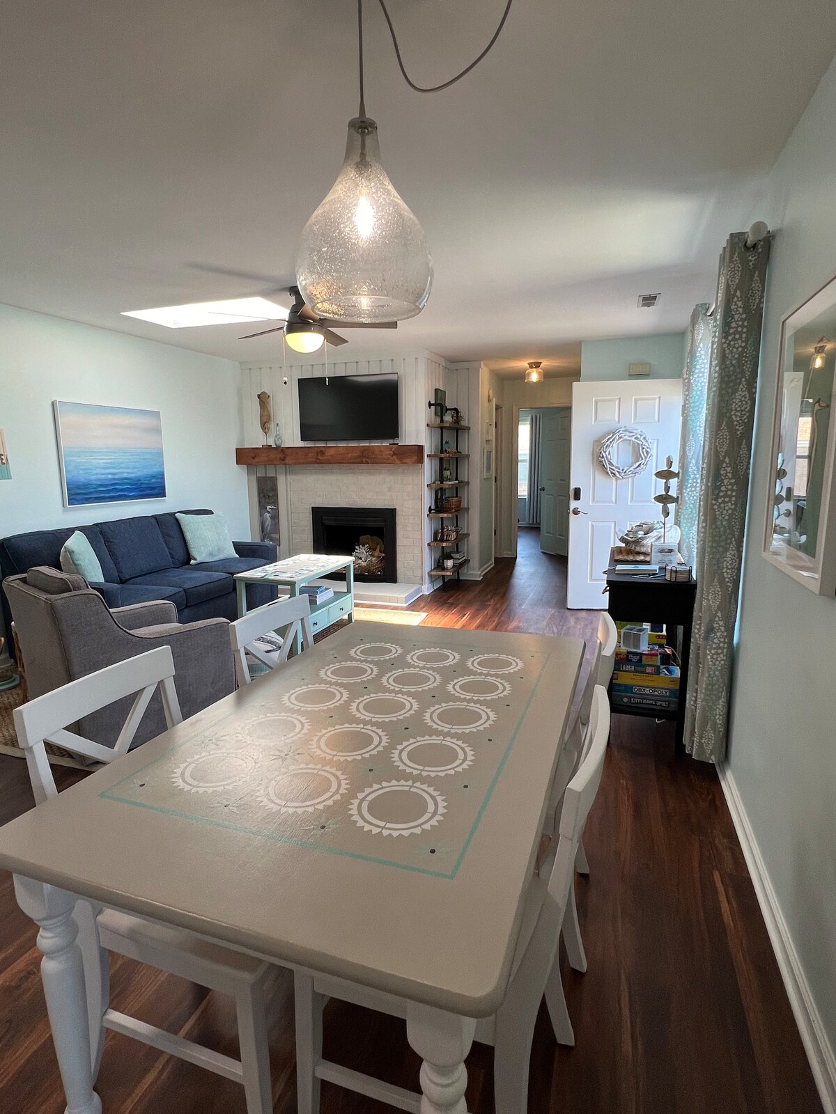 A bright living space features a light wooden dining table with decorative motifs, surrounded by white chairs. A cozy seating area is visible with a gray couch, adorned with soft cushions. Natural light floods through a skylight, enhancing the open and airy ambiance.