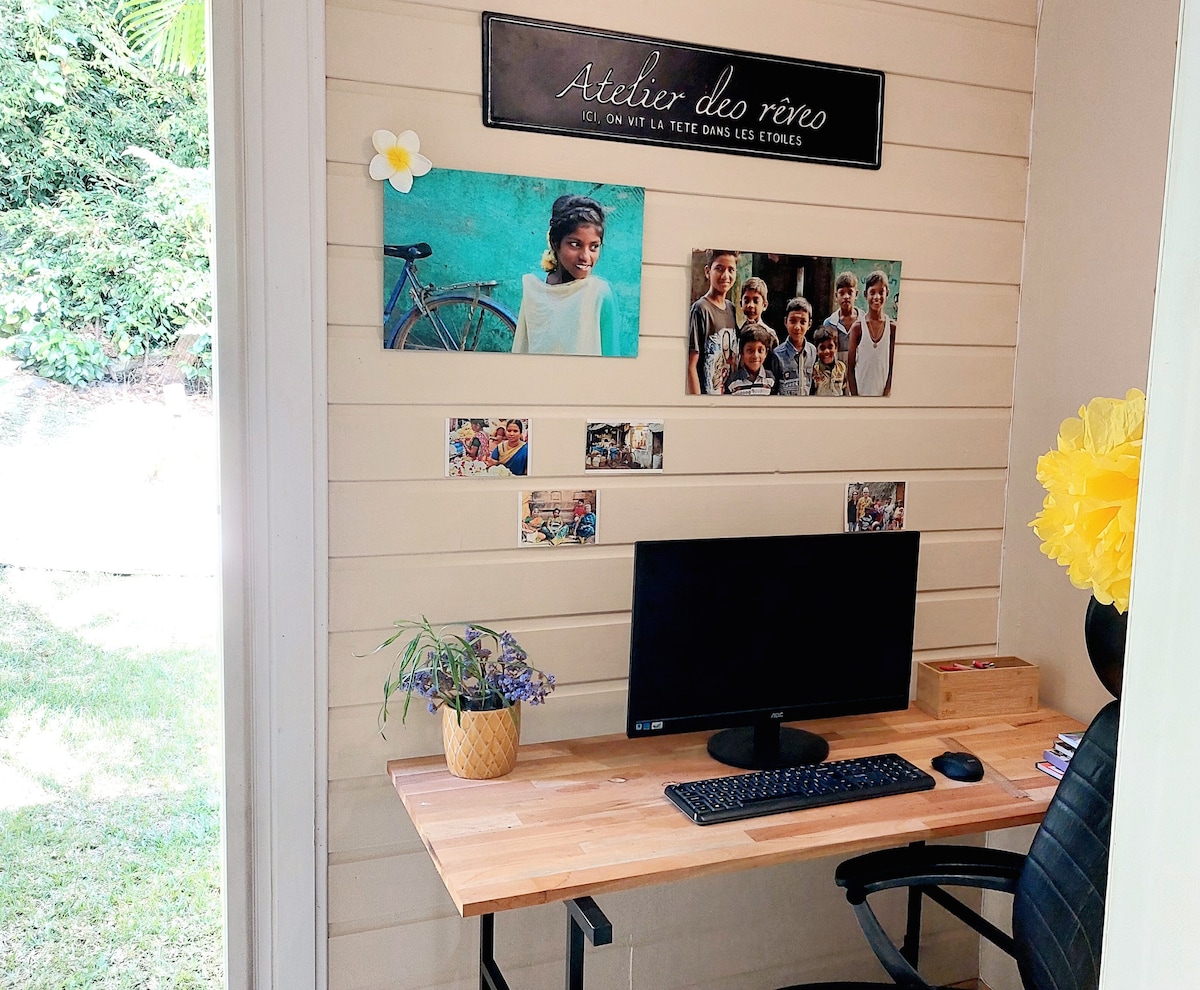 A dedicated workspace is displayed with a wooden desk and a computer monitor. Framed photographs and decorative items enhance the wall, while a potted plant adds a touch of greenery. Natural light is introduced through a nearby opening, creating an inviting environment.