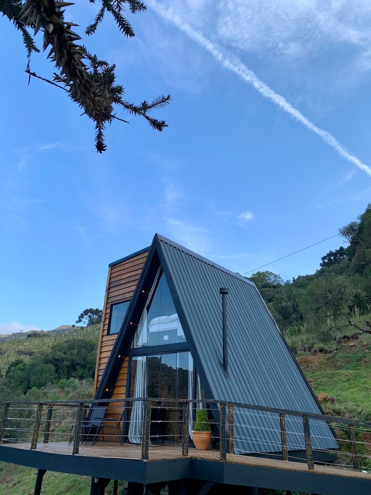 A modern A-frame cabin is set against a backdrop of lush greenery. Large windows allow natural light to flood the interior, while a balcony offers a view of the surrounding landscape. The structure combines wood and metal materials, enhancing its contemporary design.