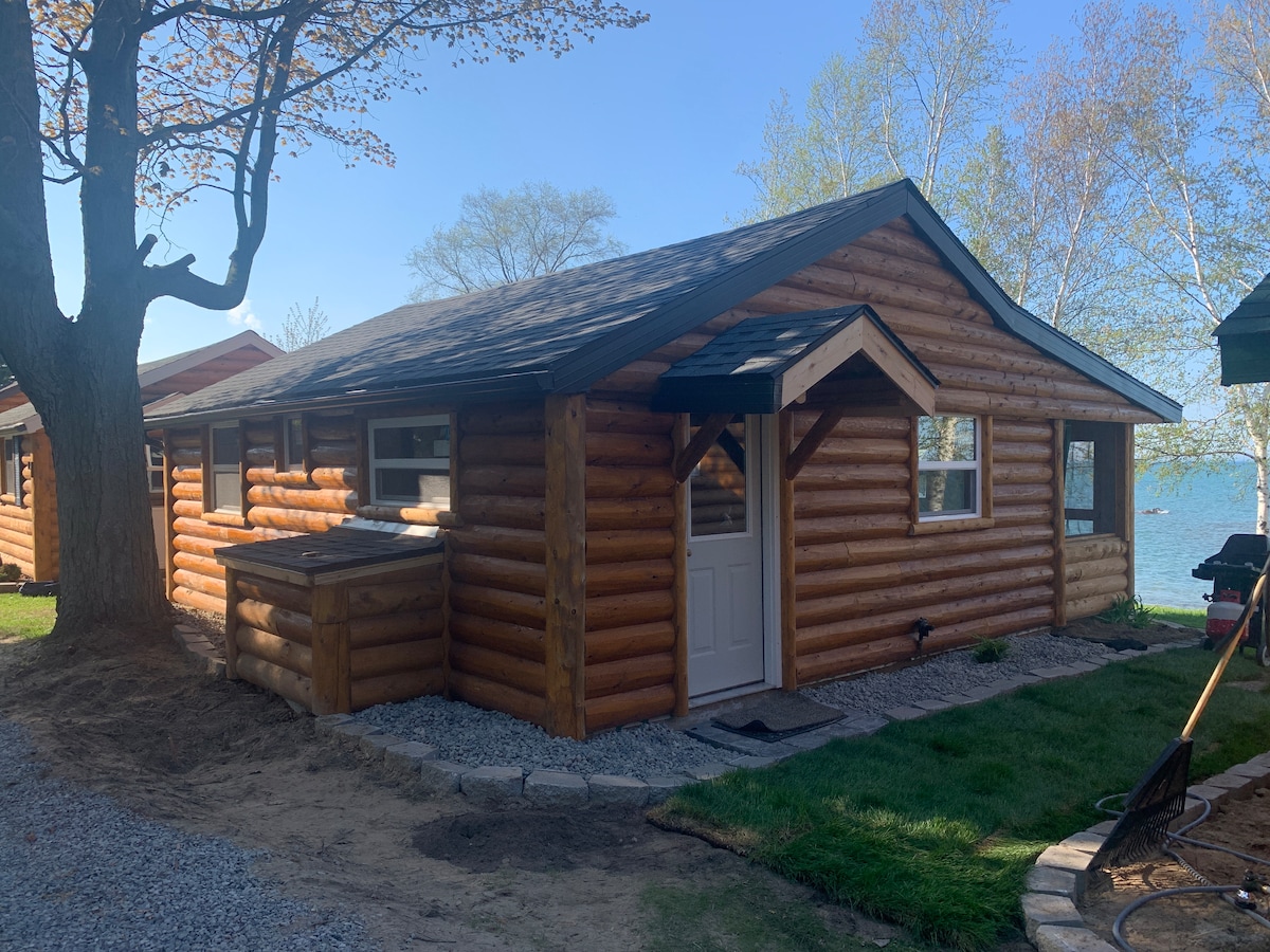 The exterior of a log cabin is shown, featuring natural wood siding and a welcoming entrance. A path of gravel leads to the door, surrounded by well-maintained grass and trees. A glimpse of the lake can be seen in the background, enhancing the cabin's scenic setting.