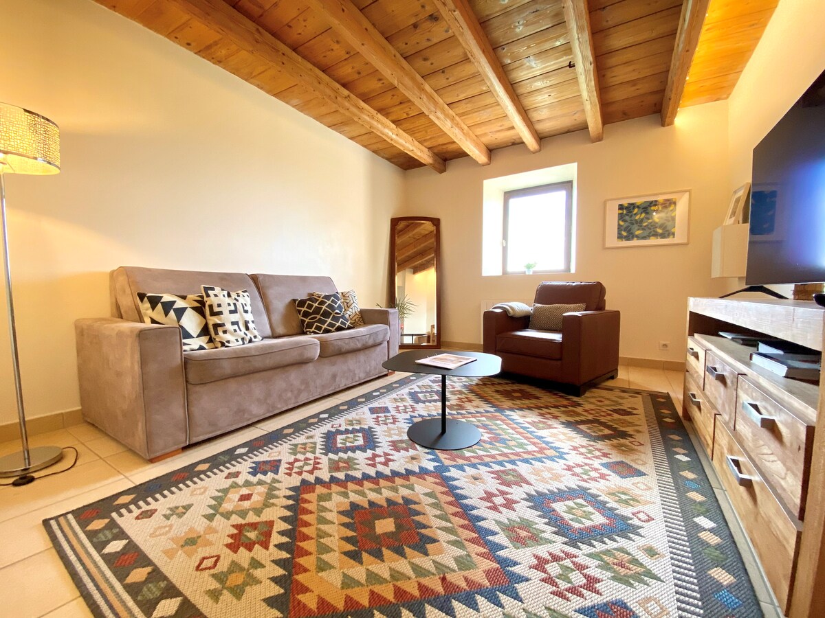 The living room features a comfortable sofa, armchair, and a low coffee table. A patterned rug is laid across the tiled floor, and natural light is provided by a window. The ceiling displays exposed wooden beams, adding warmth to the space.