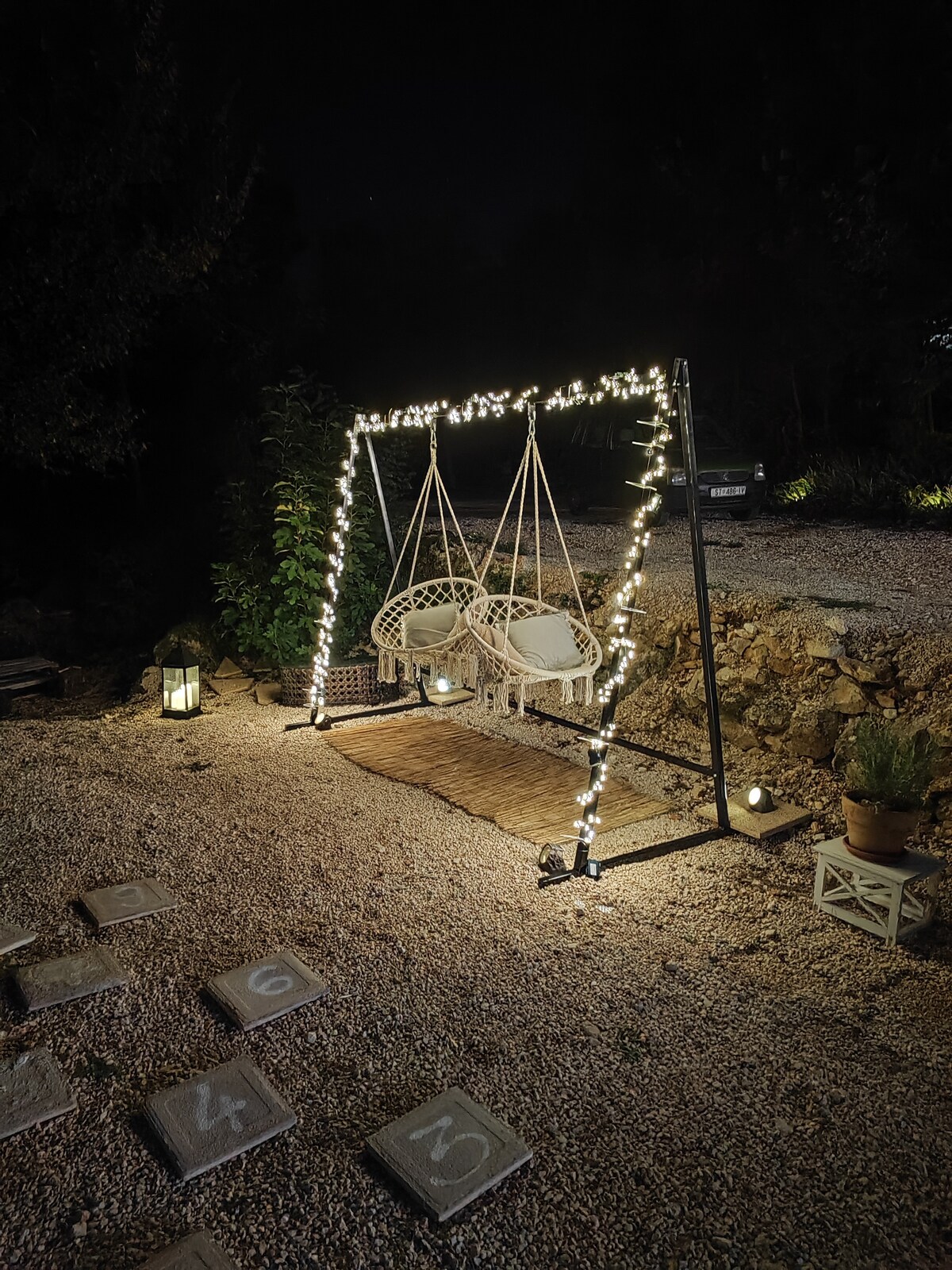 A cozy outdoor seating area features two hanging chairs, illuminated by string lights that create a warm ambiance. The gravel path leads to the chairs, surrounded by soft lighting from nearby lanterns, enhancing the serene atmosphere of the space.