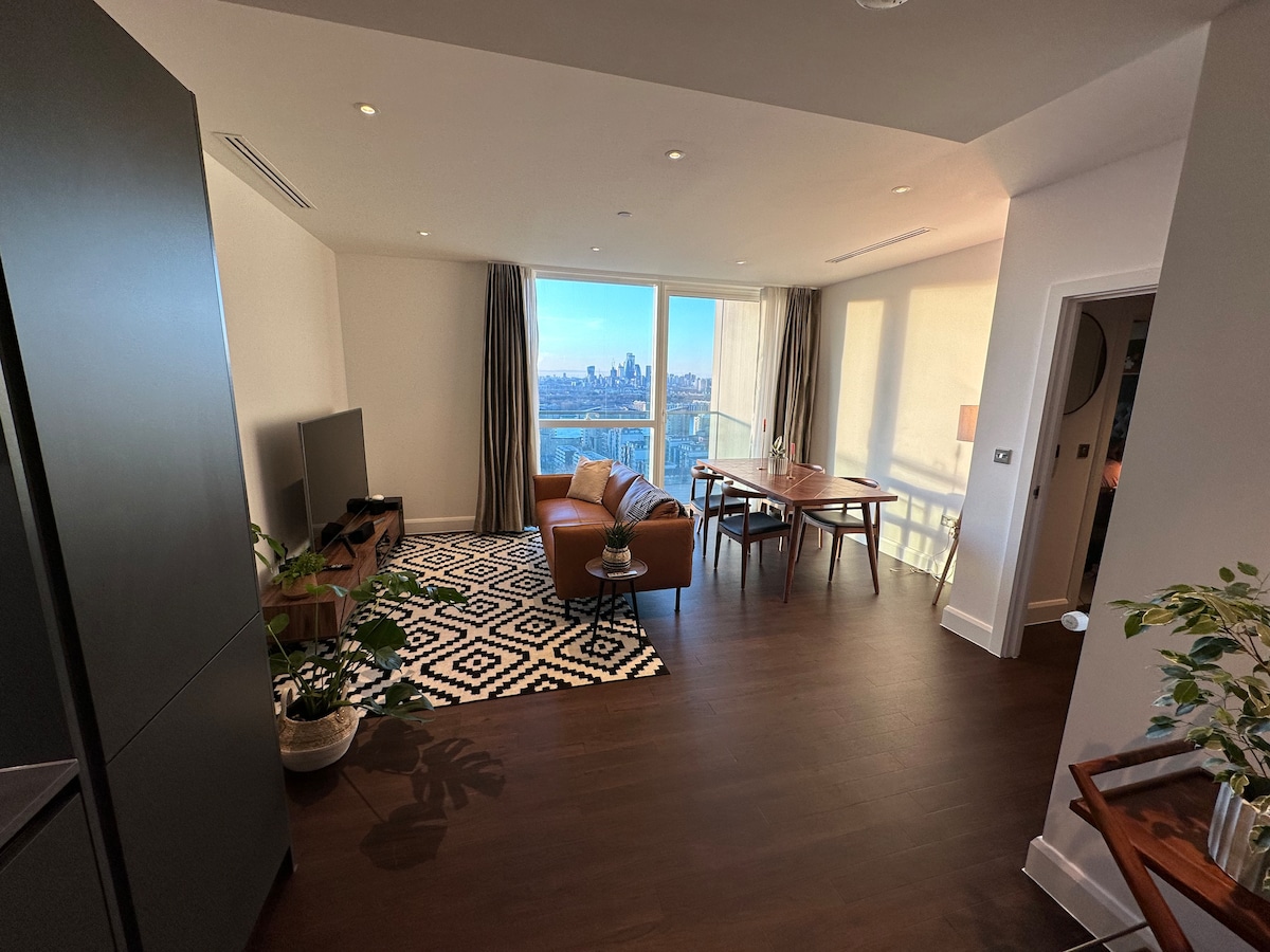 A modern living space features a leather sofa and a coffee table on a geometric-patterned rug. A television is positioned across from the seating area. A dining table sits near expansive windows, providing views of the city skyline illuminated by natural light.