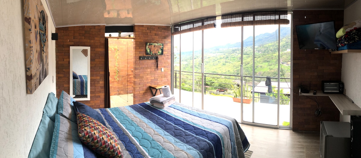 A spacious bedroom features a large bed dressed in a colorful quilt. A wall of windows provides a panoramic view of the surrounding landscape. A flat-screen TV hangs above a desk, while natural light brightens the warm-toned interior accentuated by textured brick walls.