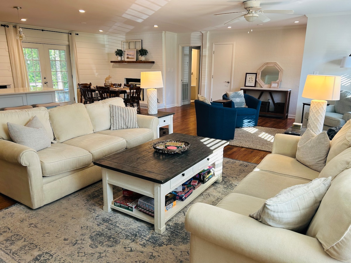 A spacious great room features two plush sofas flanking a coffee table, adorned with books and decorative items. Natural light filters through a nearby window, illuminating the warm wooden floor and accents throughout the area. A desk and chair are positioned against one wall.