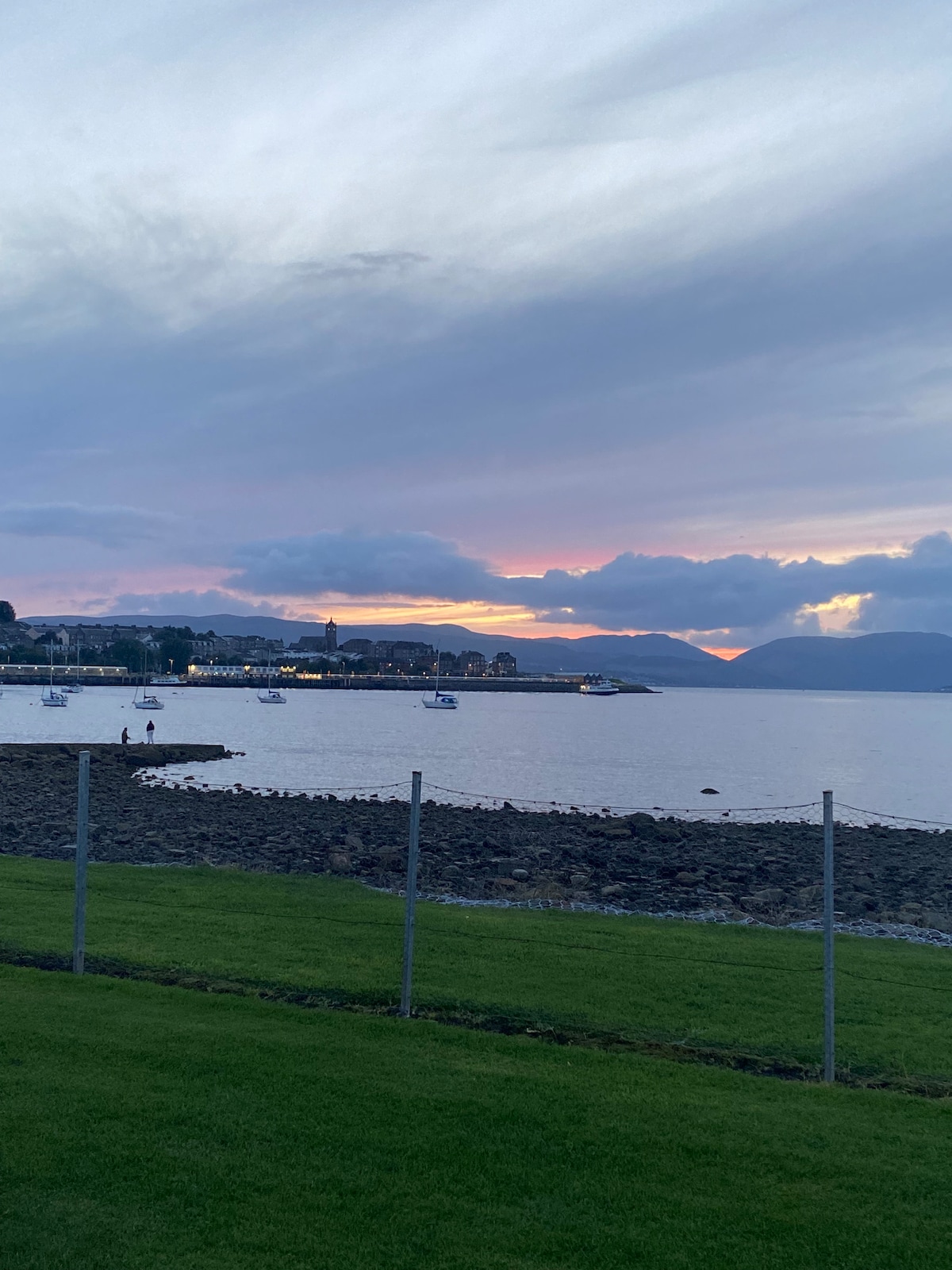A scenic waterfront view is framed by a fence, showcasing a calm bay with boats in the water. Soft colors of twilight paint the sky and reflect on the surface, while silhouettes of distant hills complete the tranquil landscape.