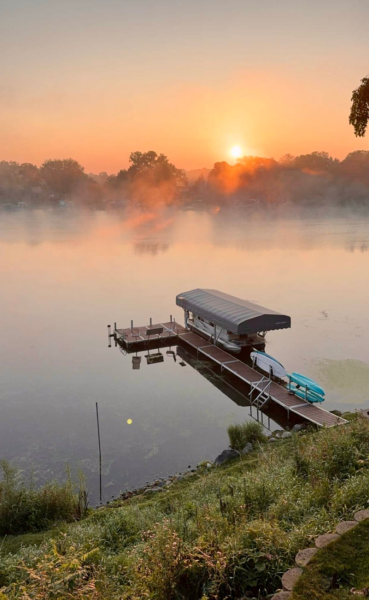A serene lakeside scene features a dock with a covered lift, accommodating a pontoon. Calm waters reflect the warm hues of a sunrise, while a gentle mist hovers above the lake. The surrounding greenery enhances the tranquil atmosphere.