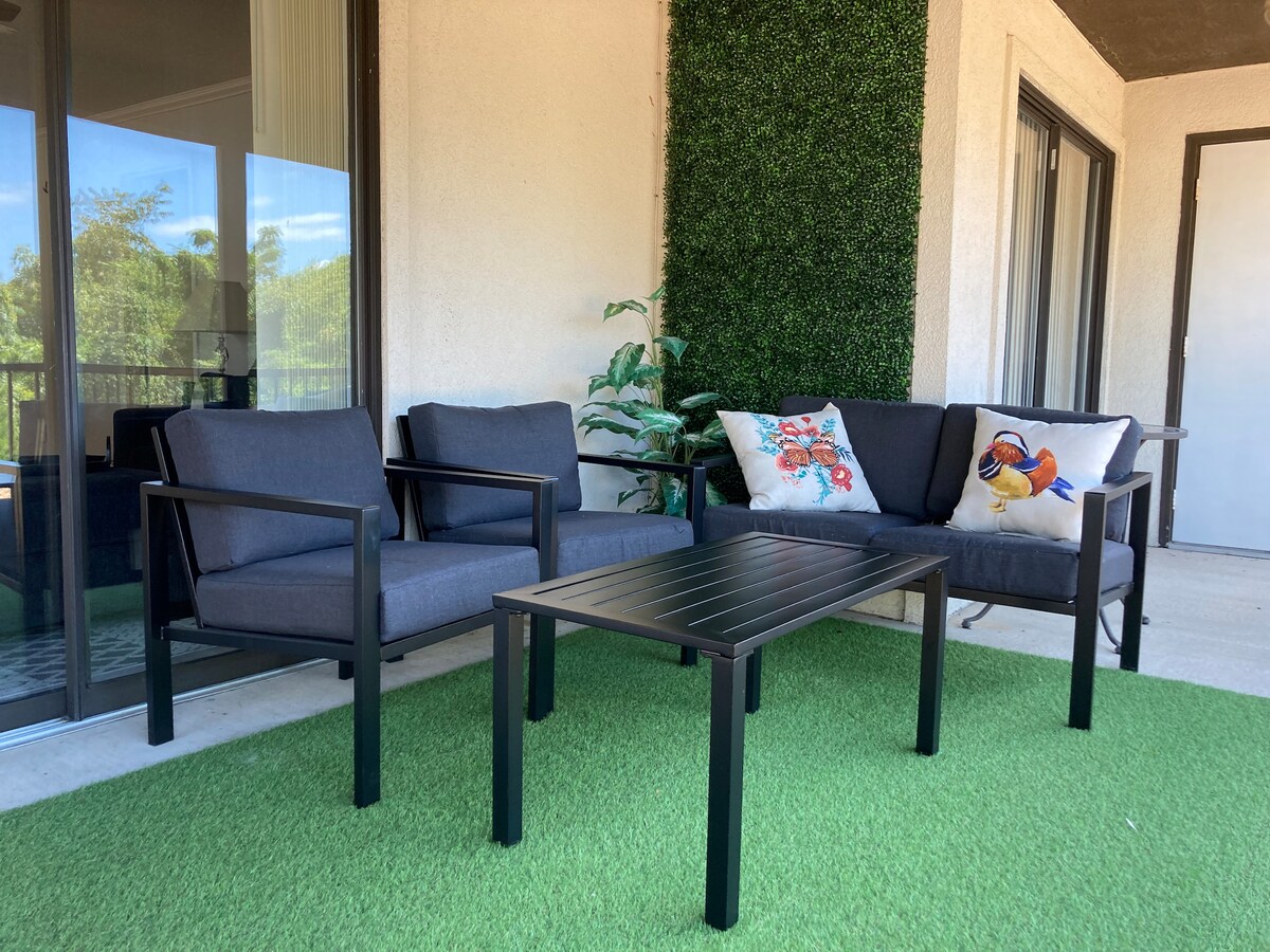 A back patio offers a relaxing seating area with two modern chairs and a sofa, accompanied by a small coffee table. Decorative pillows add a touch of color, while a lush green mat and potted plant enhance the outdoor ambiance.