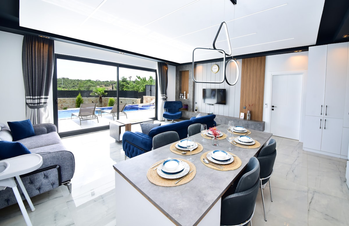 The modern living space features a dining area with a set table and a sleek kitchen counter. A cozy seating arrangement is positioned nearby, with large glass doors opening to an outdoor pool area. Natural light enhances the room's spaciousness, complemented by stylish decor.