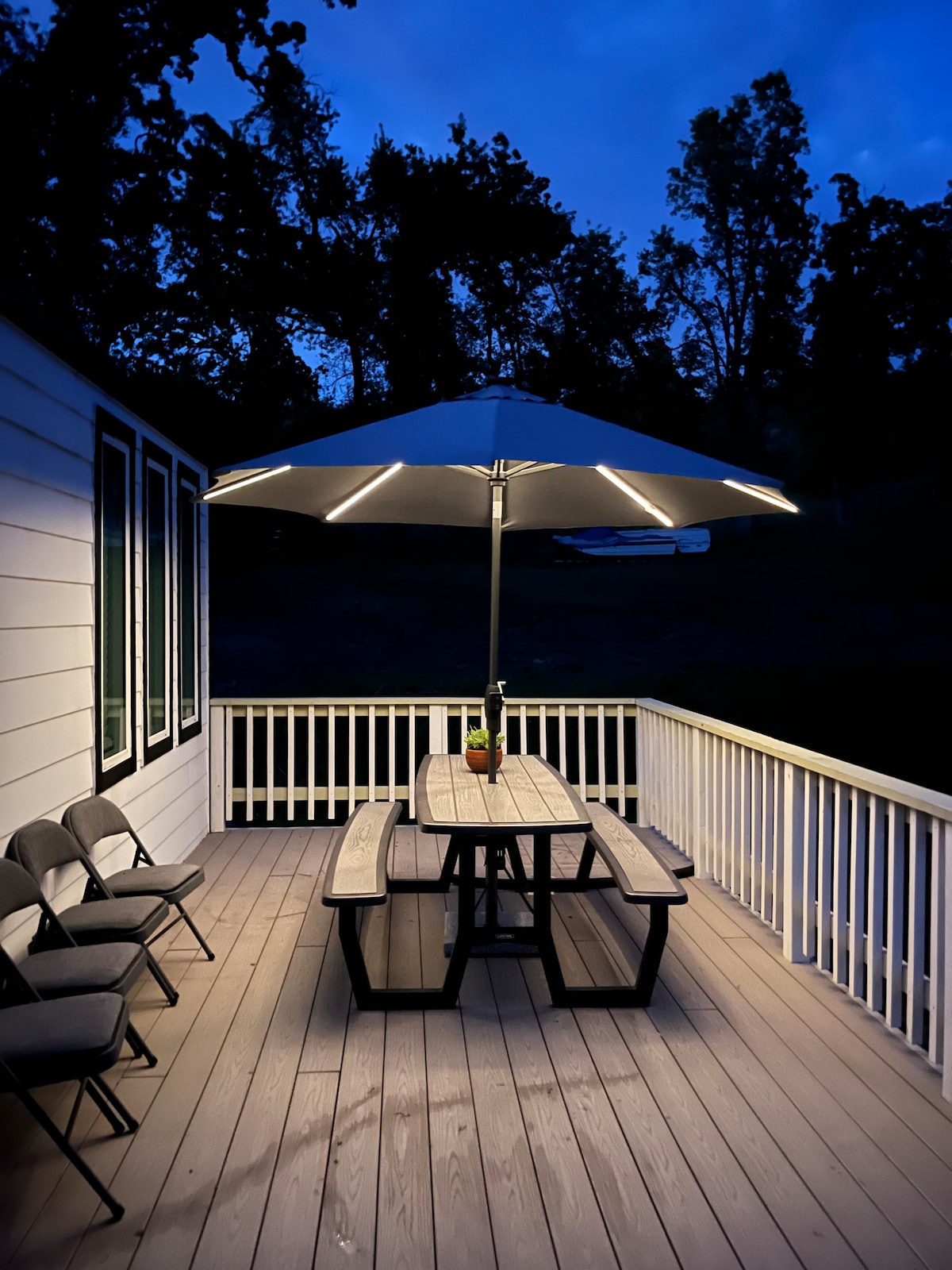 A cozy outdoor dining area is illuminated under a large umbrella, showcasing a central picnic table surrounded by several folding chairs. The decking is made of light wood, and the surrounding space features trees in the background, creating a serene outdoor ambiance.