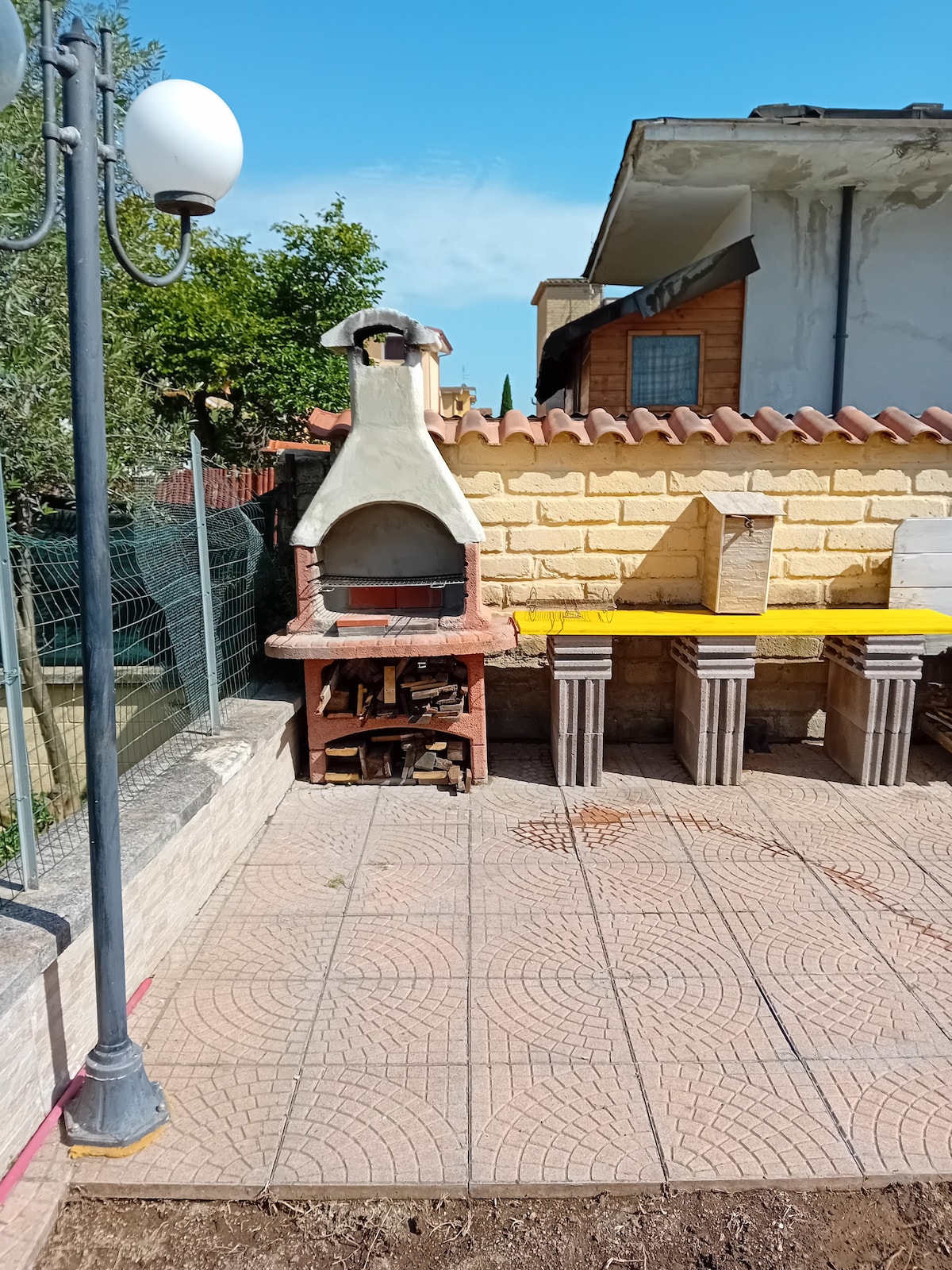 An outdoor space features a stone barbecue grill, complemented by wooden logs stacked nearby. Two rustic tables with concrete bases and yellow tabletops provide ample seating. The area is paved with patterned tiles and is set against a backdrop of greenery and a clear blue sky.