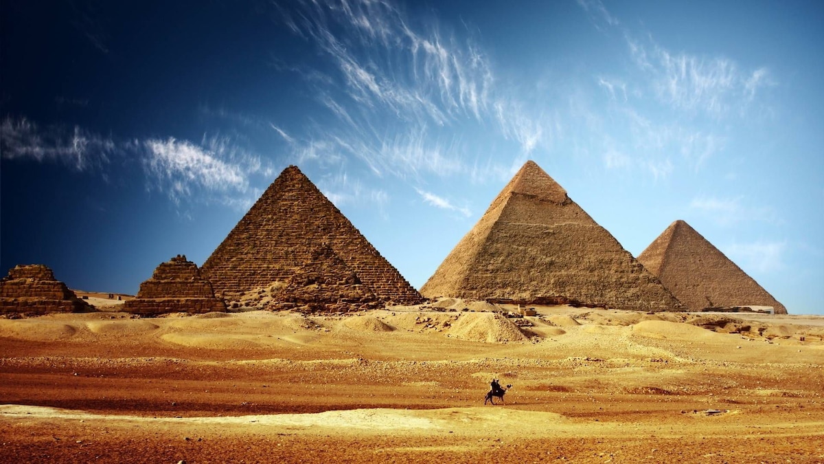 A wide view of the iconic pyramids set against a clear blue sky, with minimal clouds. The sandy landscape surrounds the structures, highlighting their geometric shapes and ancient textures. A figure on horseback can be seen in the foreground, adding a sense of scale to the monumental scene.