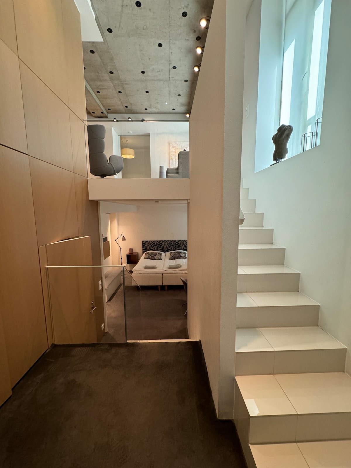 A modern interior is captured from the lower level, showcasing a staircase that leads to an upper area. Smooth white walls and polished tiles create a clean look. In the background, a spacious bed is slightly visible through short walls, with large windows allowing natural light to enter.