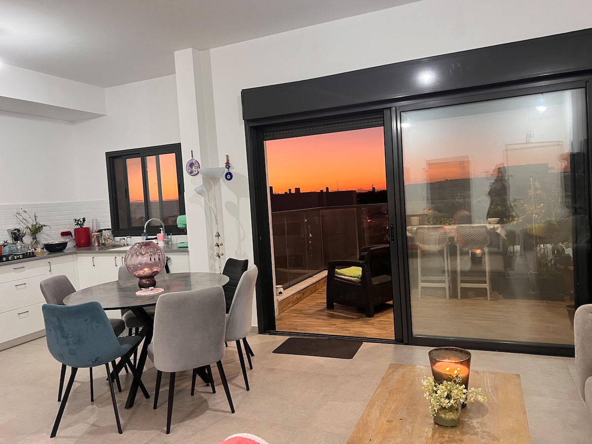 A modern dining area features a round table with stylish chairs, bathed in soft evening light. Large glass doors open to a balcony, offering a view of a colorful sunset. A cozy candlescape is centered on a wooden surface, enhancing the ambiance.
