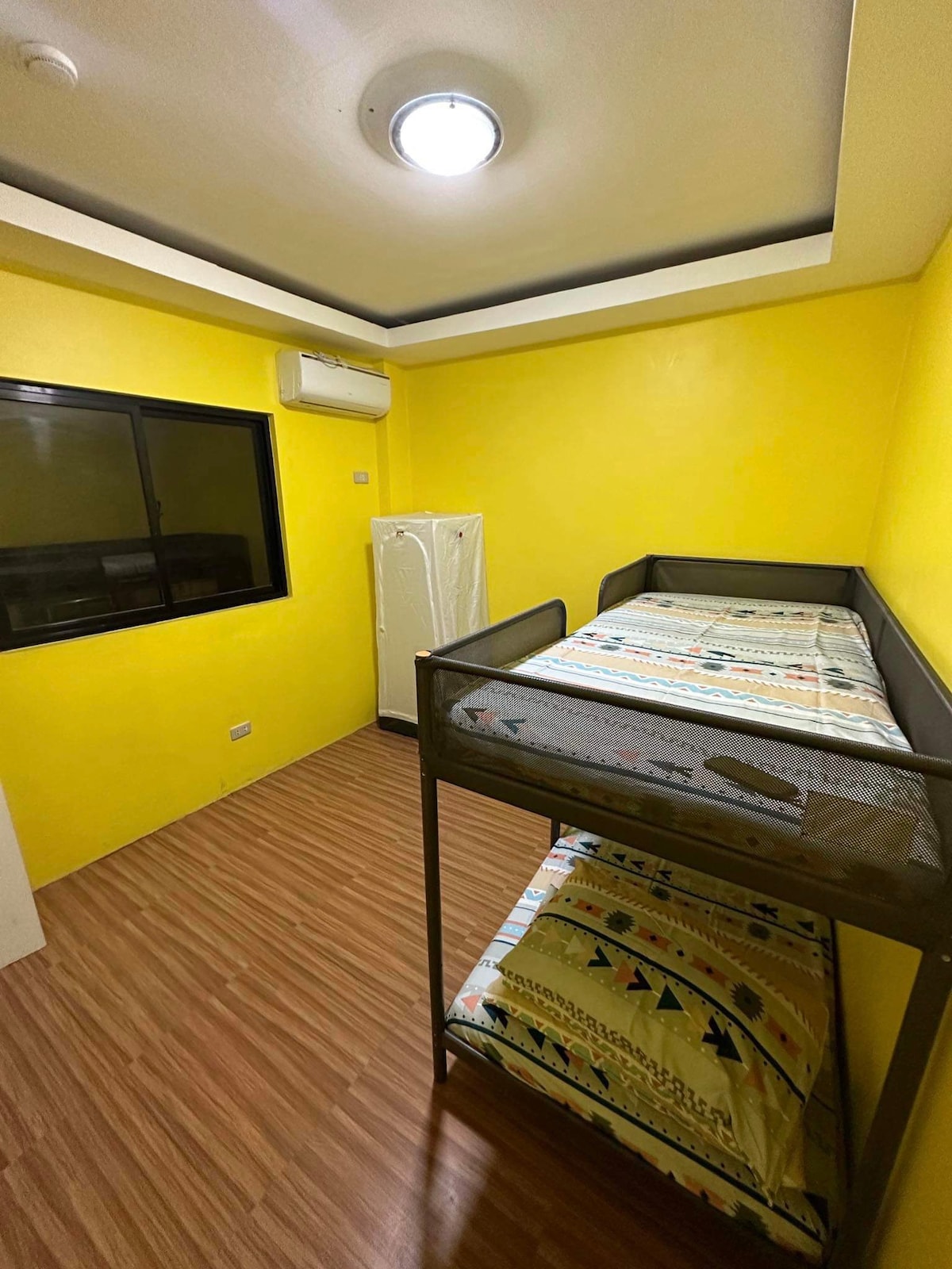 A small room is presented with vibrant yellow walls and a light wooden floor. A metal bunk bed is placed against one wall, featuring a patterned mattress. A window allows natural light to enter, and a refrigerator is visible nearby.