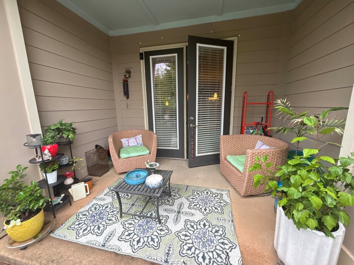 An inviting entryway features two wicker chairs with green cushions positioned beside a decorative table. A patterned rug lies beneath, complemented by potted plants and a whimsical decoration on the wall. A ladder leans against the wall, enhancing the welcoming atmosphere.