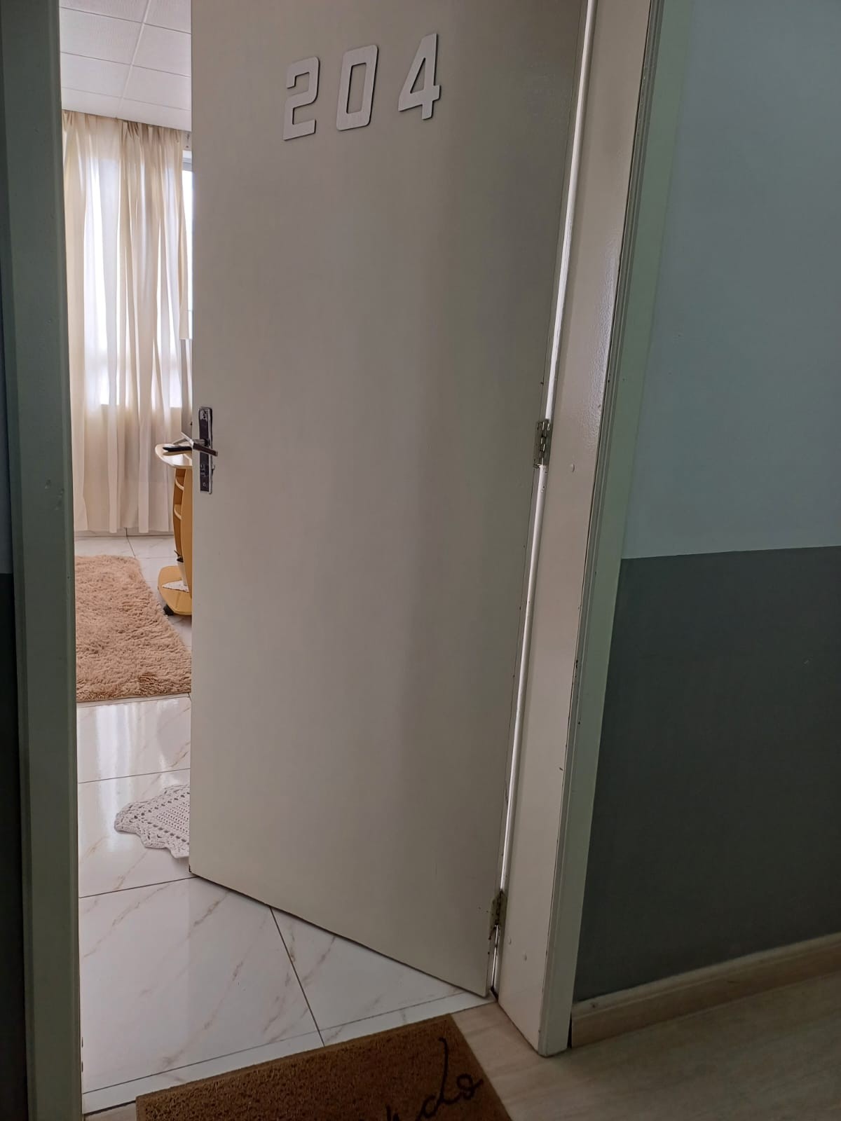 A white door marked with number 204 is slightly ajar, leading to a well-lit interior space. Soft, neutral-toned curtains drape beside a window, and a plush, light-colored rug is visible on the tiled floor. The inviting ambiance suggests a cozy atmosphere.