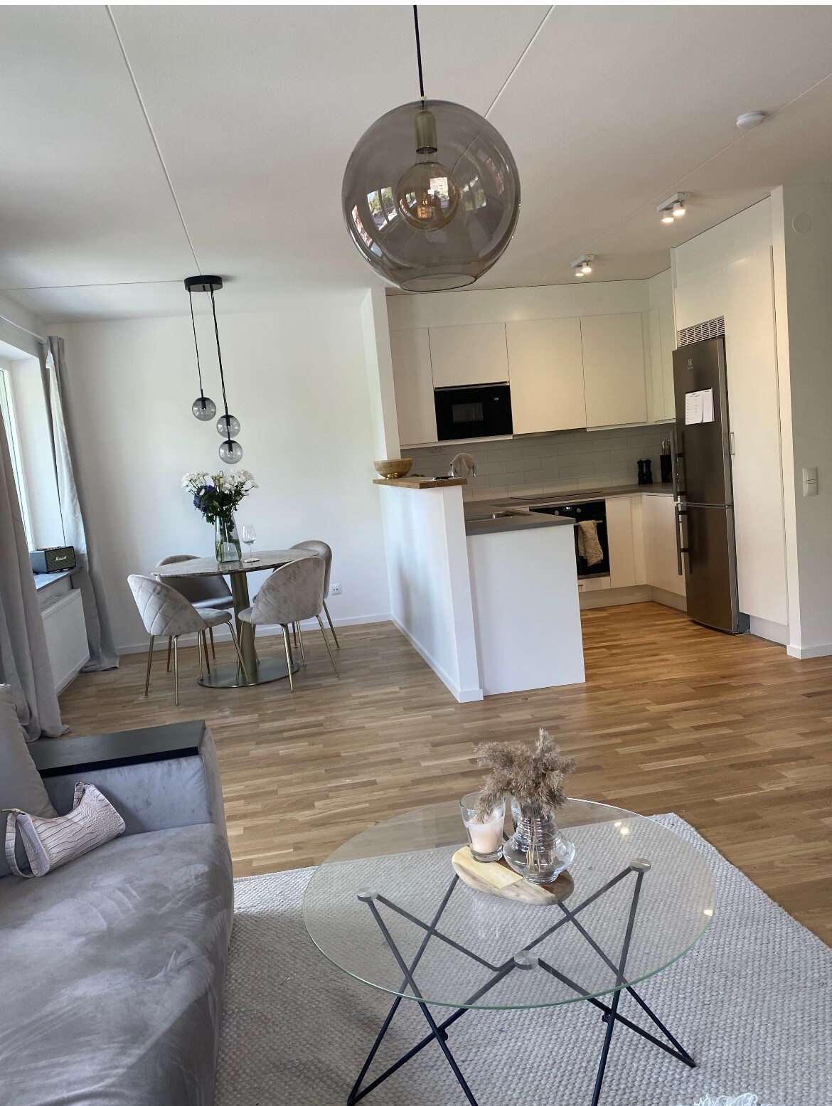 A modern living area features a light-colored sofa and a glass coffee table with decorative items. The dining space includes a round table surrounded by upholstered chairs, with a well-equipped kitchen visible in the background. Natural light streams through large windows.