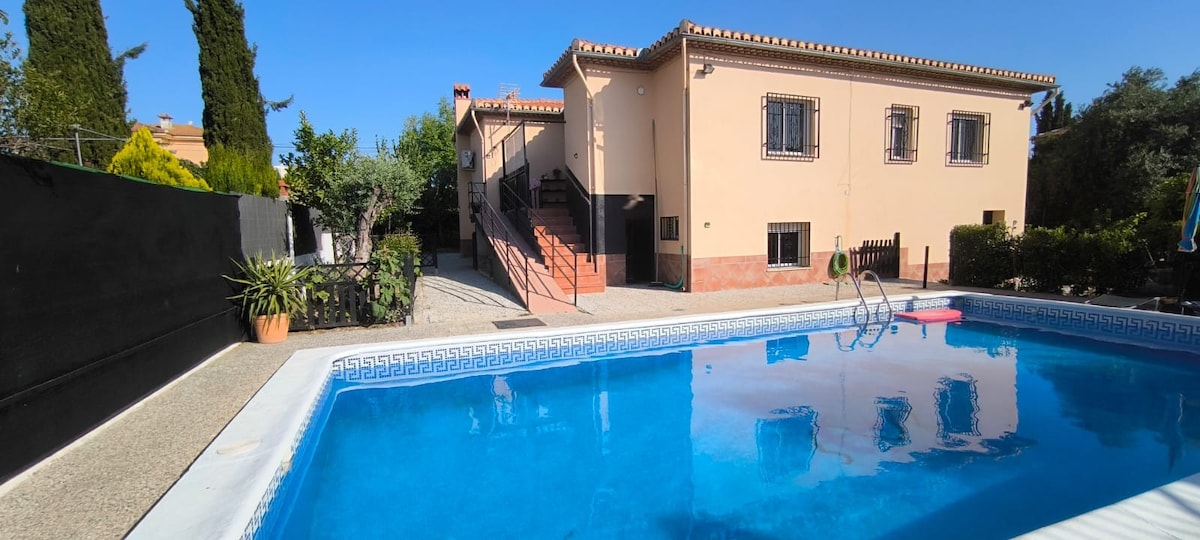 A spacious outdoor area features a clear swimming pool reflecting the sunny sky. The pool is surrounded by greenery and a pathway leading to the two-story house, which displays classic architectural details and several windows.