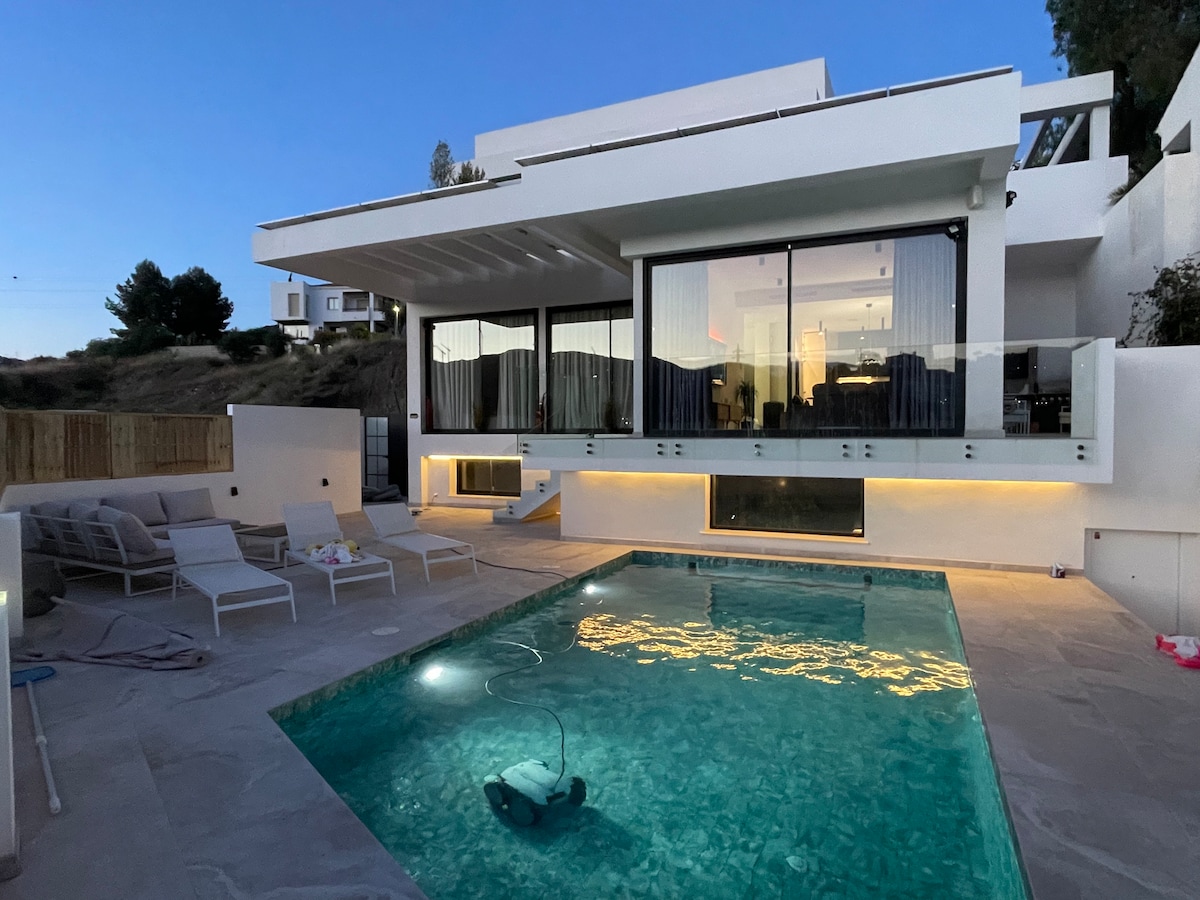 A modern villa showcases a spacious outdoor area featuring a swimming pool with illuminated water. Large glass doors invite natural light into the living space above, while lounge chairs line the poolside. Soft lighting enhances the tranquil ambiance during the evening.