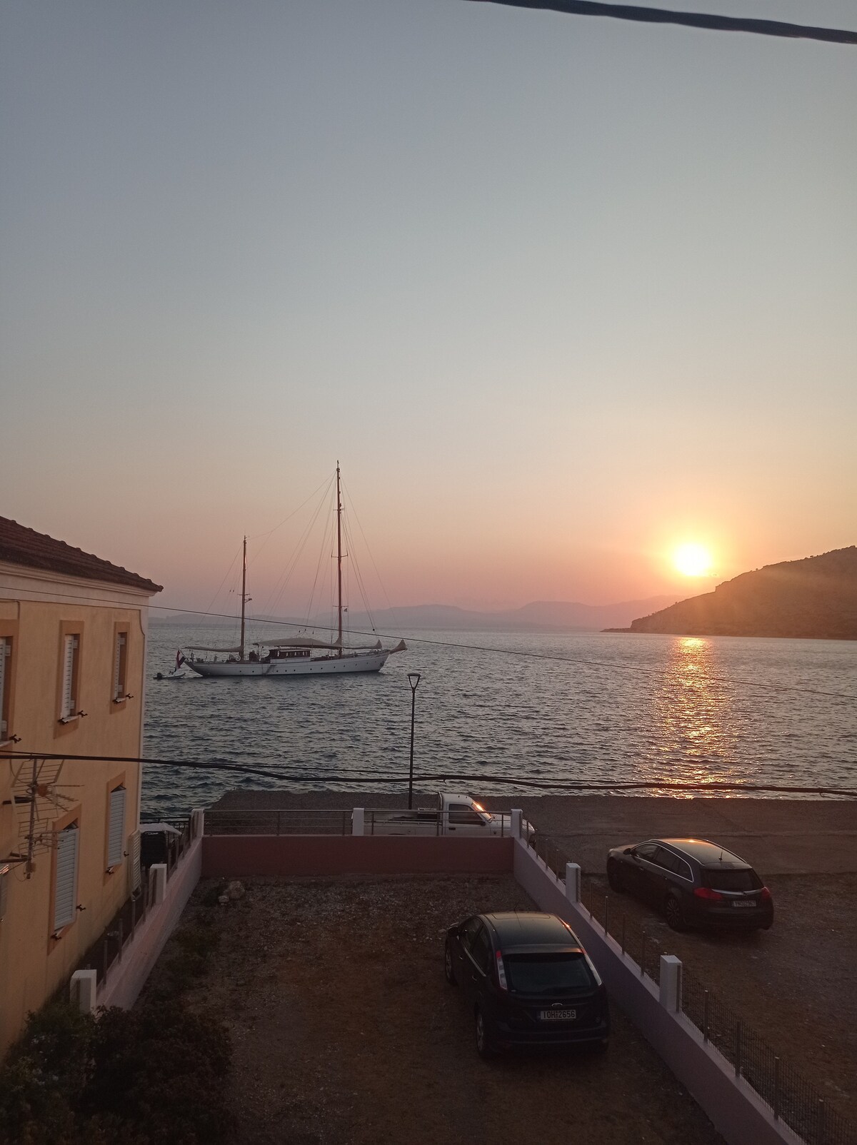 A sailboat is seen gently floating on calm waters during sunset. The sun is positioned near the horizon, casting a warm glow over the scene. A sandy area is located in the foreground with parked cars and a light-colored building in the background.