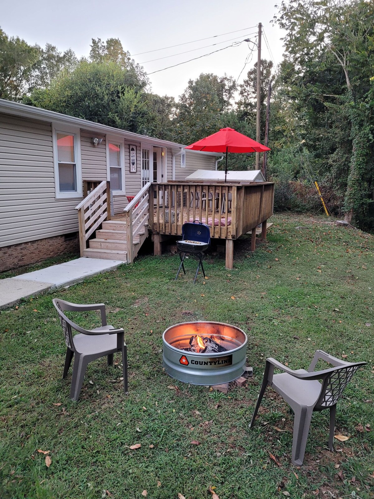 A cozy outdoor space features a fire pit surrounded by three chairs on a grassy area. A wooden deck is accessible via stairs. An umbrella provides shade over the deck, contributing to the inviting atmosphere of the backyard.