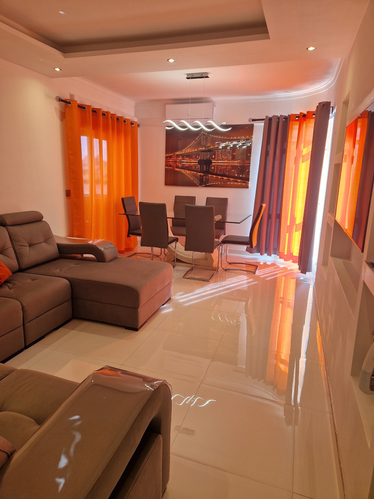 A living room area is presented, featuring a large, L-shaped sofa in a neutral tone. A glass dining table surrounded by six chairs sits nearby. Bright orange curtains frame the windows, allowing natural light to illuminate the glossy tiled floor.