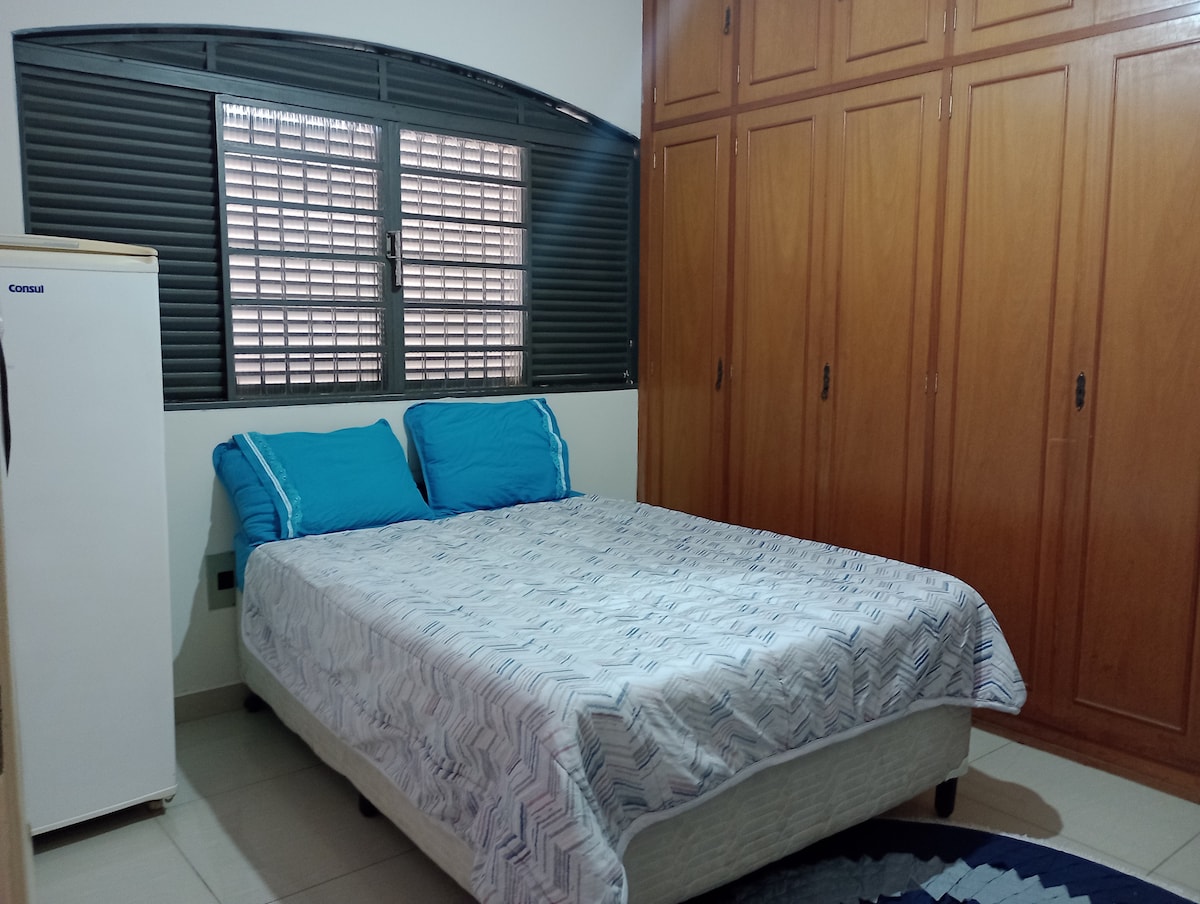 A furnished bedroom features a spacious bed with blue accent pillows and a patterned bedspread. A white refrigerator stands nearby, and a large window with horizontal blinds allows natural light. Ample wooden cabinetry lines the wall, providing storage options.