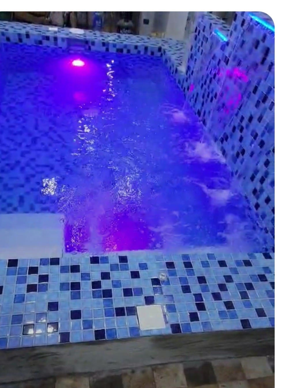 A dipping pool is presented, featuring a mosaic of blue tiles with accents of white and darker shades. Underwater lighting casts a soft purple glow, creating a soothing ambiance. Bubbles emerge from the pool, suggesting active water features within the space.