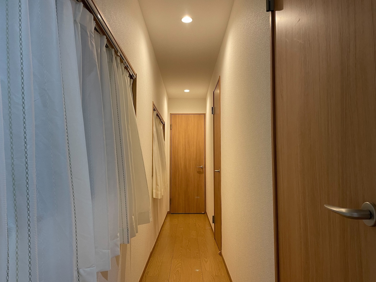 A well-lit hallway is lined with light wood flooring and white walls. Soft curtains hang beside the doorways, creating a clean and welcoming appearance. Doors are positioned at either end of the corridor, emphasizing a sense of openness and accessibility.