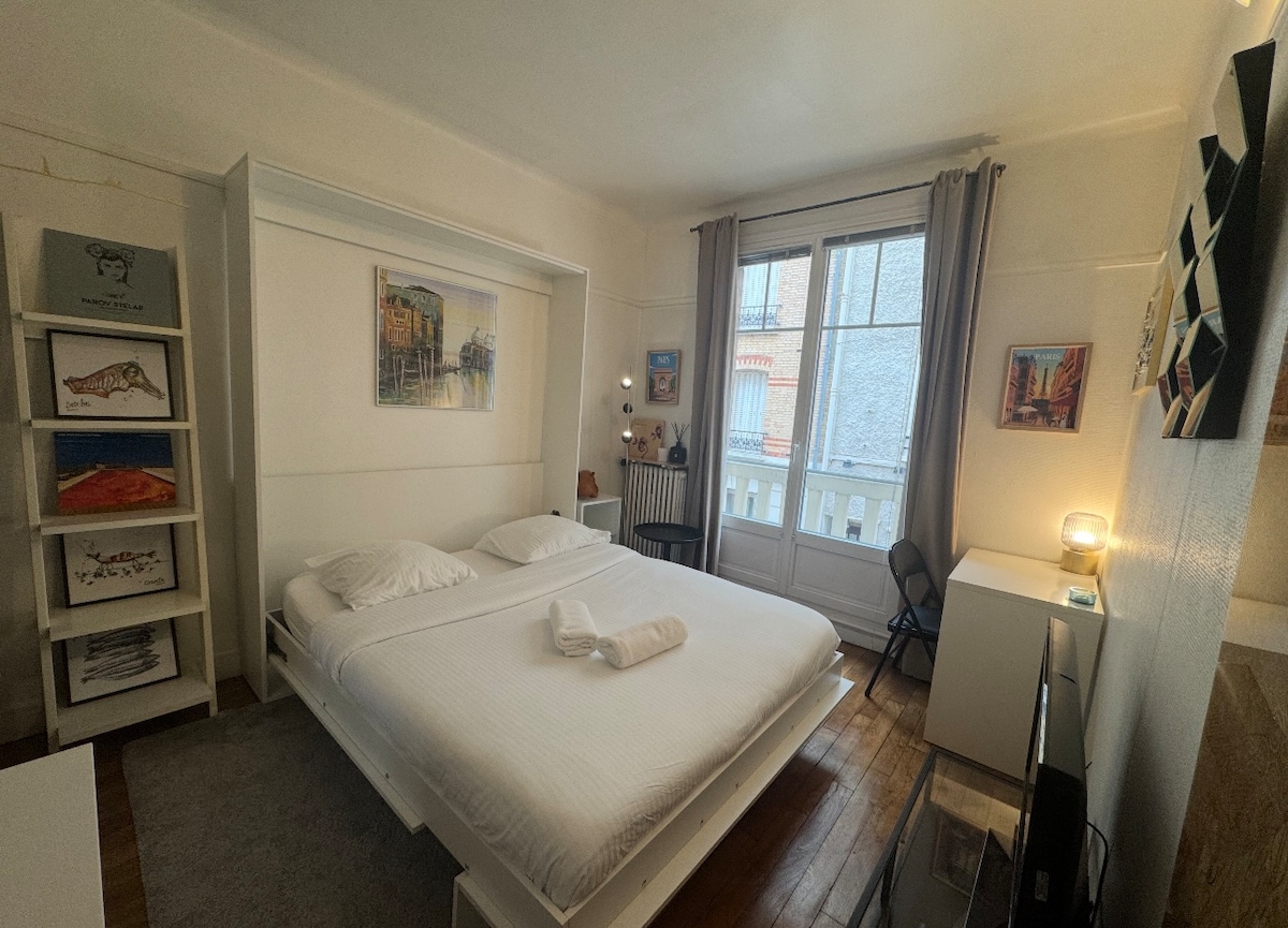A well-lit studio features a spacious bed with white linens, complemented by artwork on the walls. A small desk is positioned beside a window, which is dressed with light curtains. There is a cozy chair and shelving displaying various items in the background.