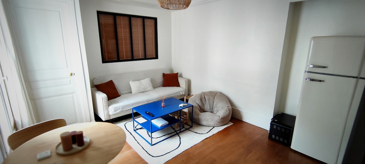 A bright living area features a comfortable white sofa with orange cushions, paired with a blue coffee table and a soft bean bag. Natural light enters through a window with wooden blinds. A round dining table is set nearby, and a vintage refrigerator adds charm to the space.