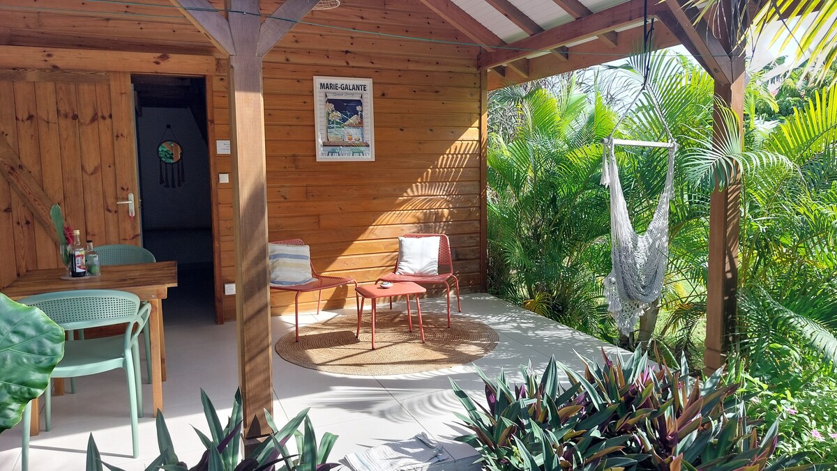 The outdoor seating area is surrounded by lush greenery, featuring a small table with two chairs and a hammock. A welcoming, natural wood texture lines the structure, complemented by a vibrant rug beneath. A tasteful piece of wall art adds character to the space.