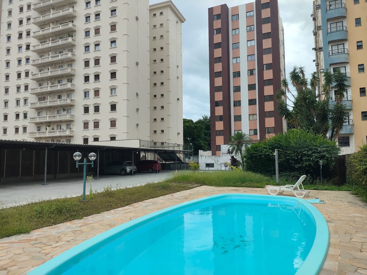 A turquoise swimming pool is visible, surrounded by a stony patio. Tall buildings stand in the background, with a mix of architectural styles. Lush greenery borders the area, providing a sense of openness and tranquility.