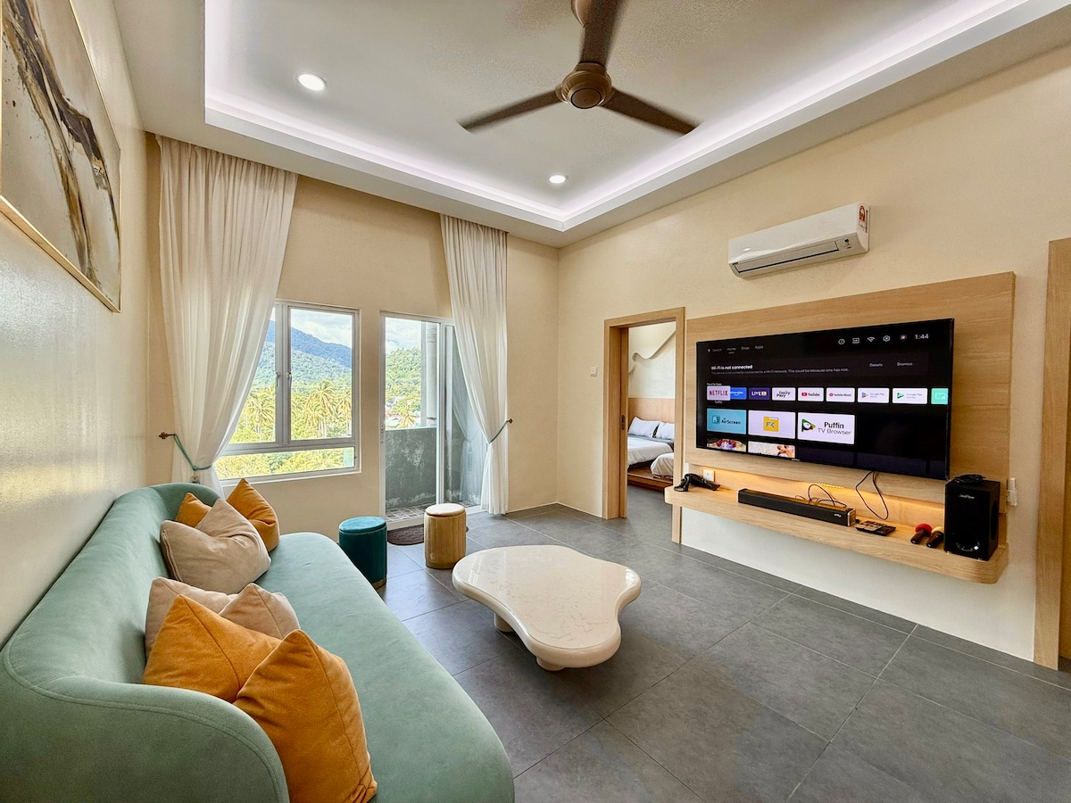 A modern living area features a soft green sofa adorned with accent pillows. A light, organic-shaped coffee table rests on the gray tile floor. The wall-mounted TV displays various apps, while large windows invite natural light and offer views of the surrounding landscape.