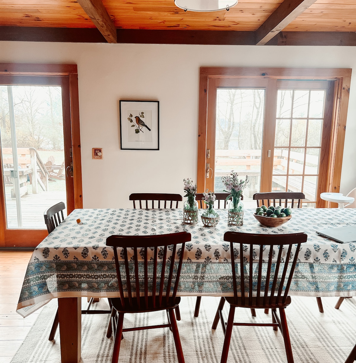 A dining area is highlighted by a large, patterned tablecloth covering a rustic wooden table surrounded by dark wooden chairs. French doors lead to an outdoor deck, and a framed bird illustration is displayed on the wall. Centerpieces of vases and a bowl enhance the table.