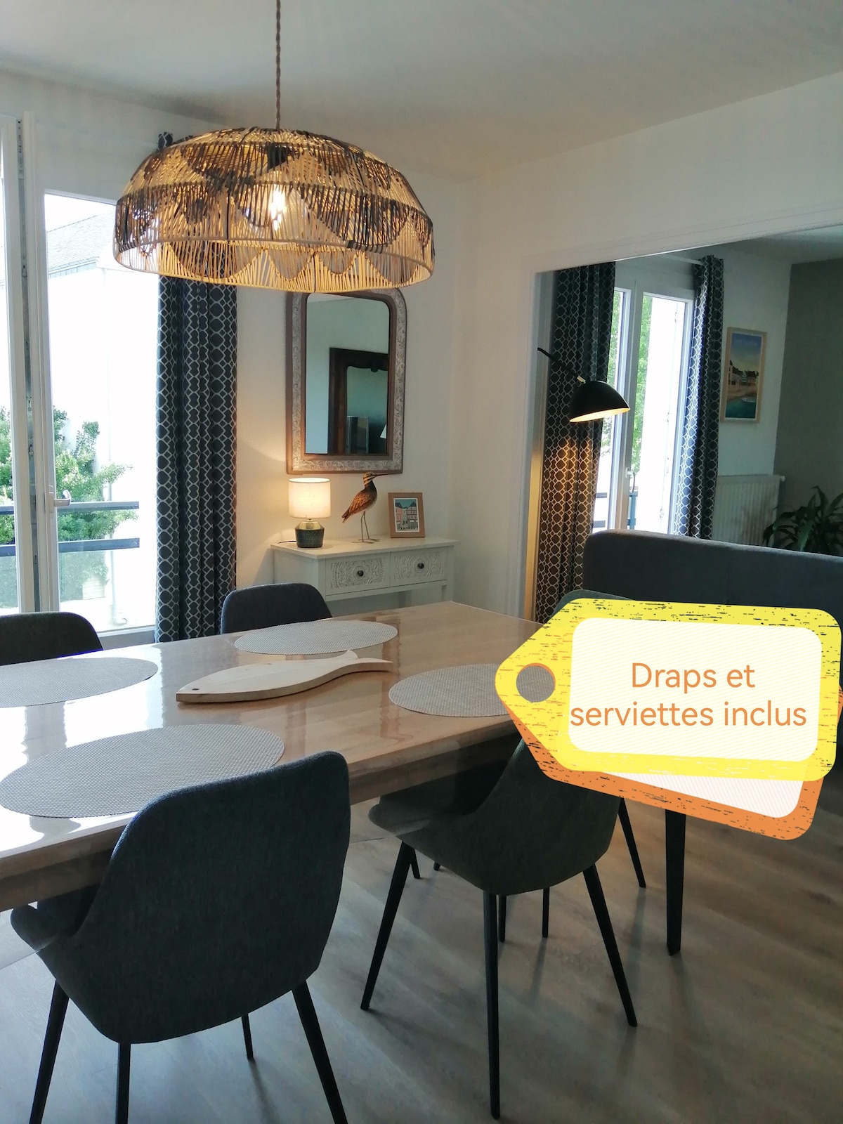 A dining area is showcased with a wooden table surrounded by six grey upholstered chairs. A stylish pendant light hangs above, complemented by a mirror and decorative items on a sideboard. Large windows allow natural light to brighten the space.